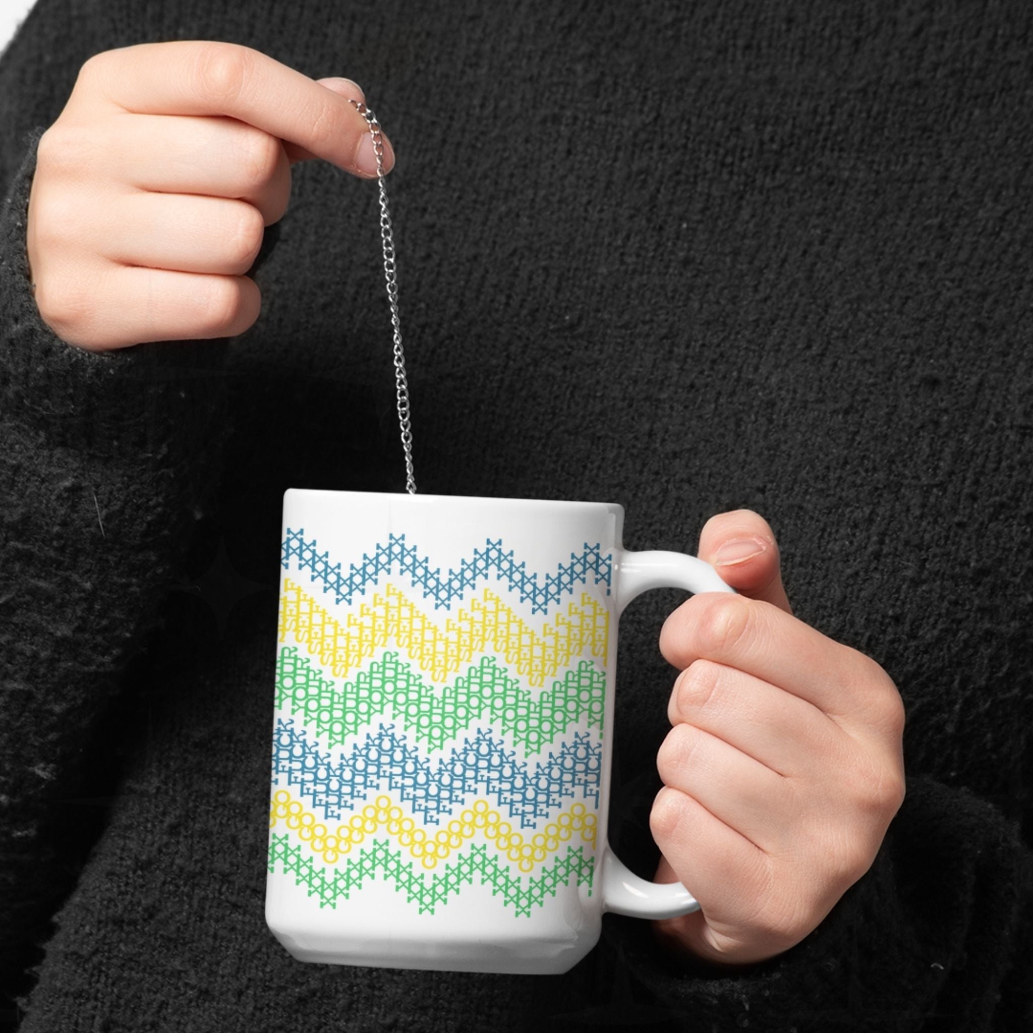 Hands holding Go Fuck Yourself cross stitch ceramic mug with tea infuser, bright teal yellow green chevron pattern