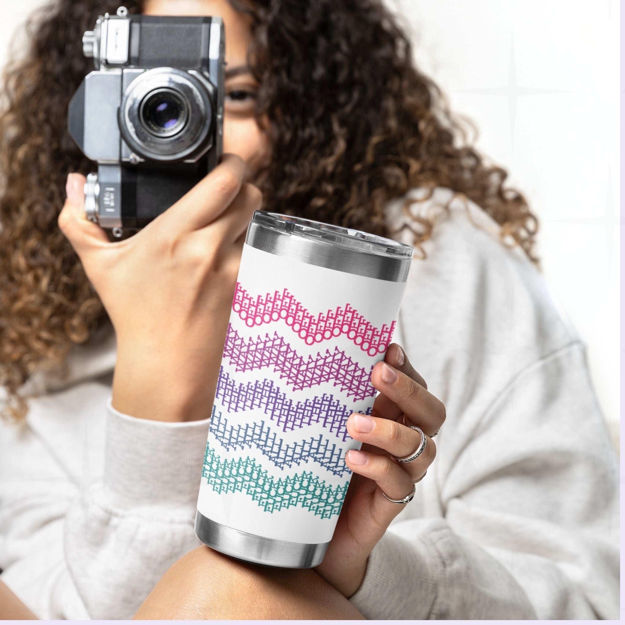 Woman with curly hair holding bright stripe hidden profanity tumbler and vintage camera showing pink purple teal cross stitch pattern