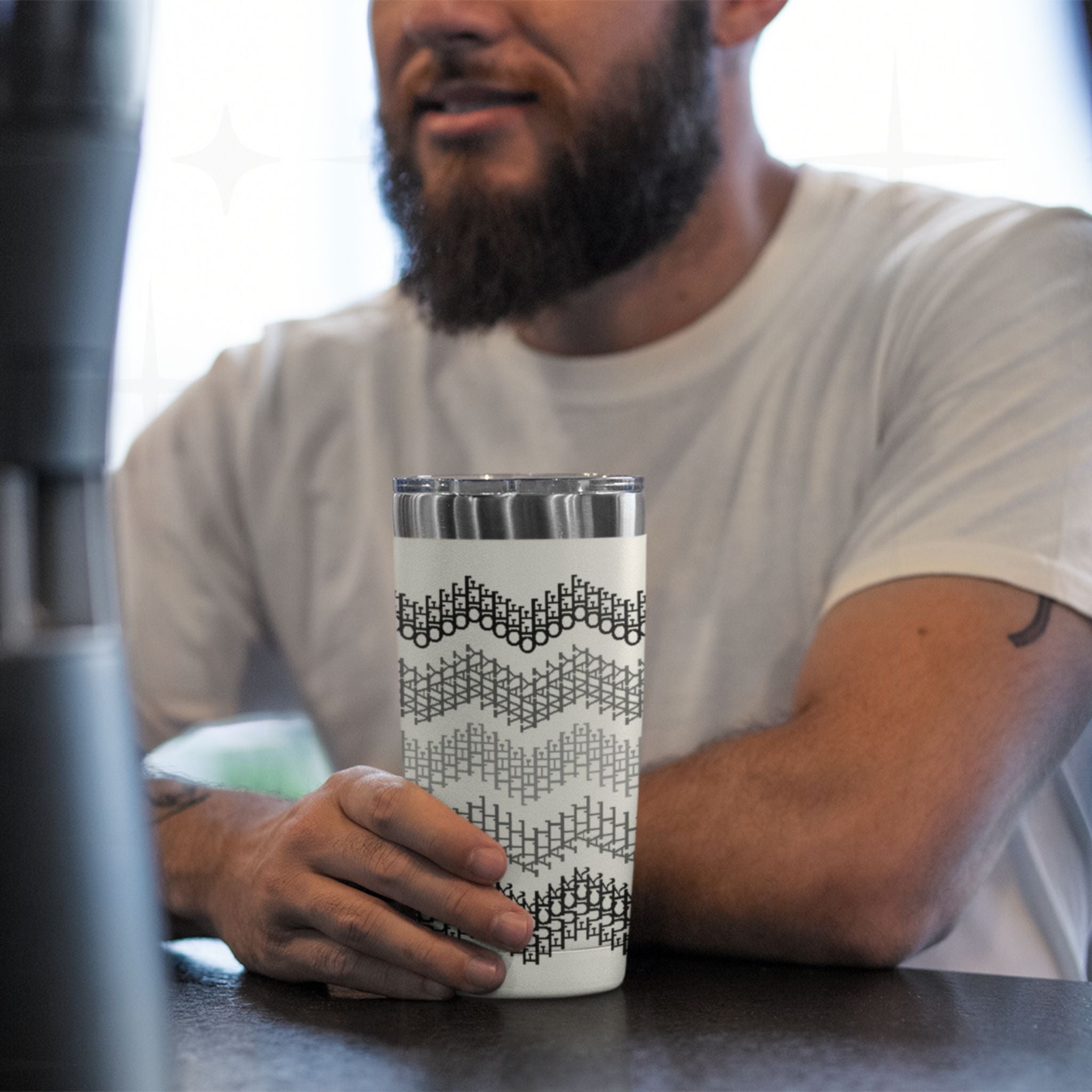Bearded man sitting at counter holding gray stripe hidden profanity tumbler with black and gray cross stitch chevron pattern