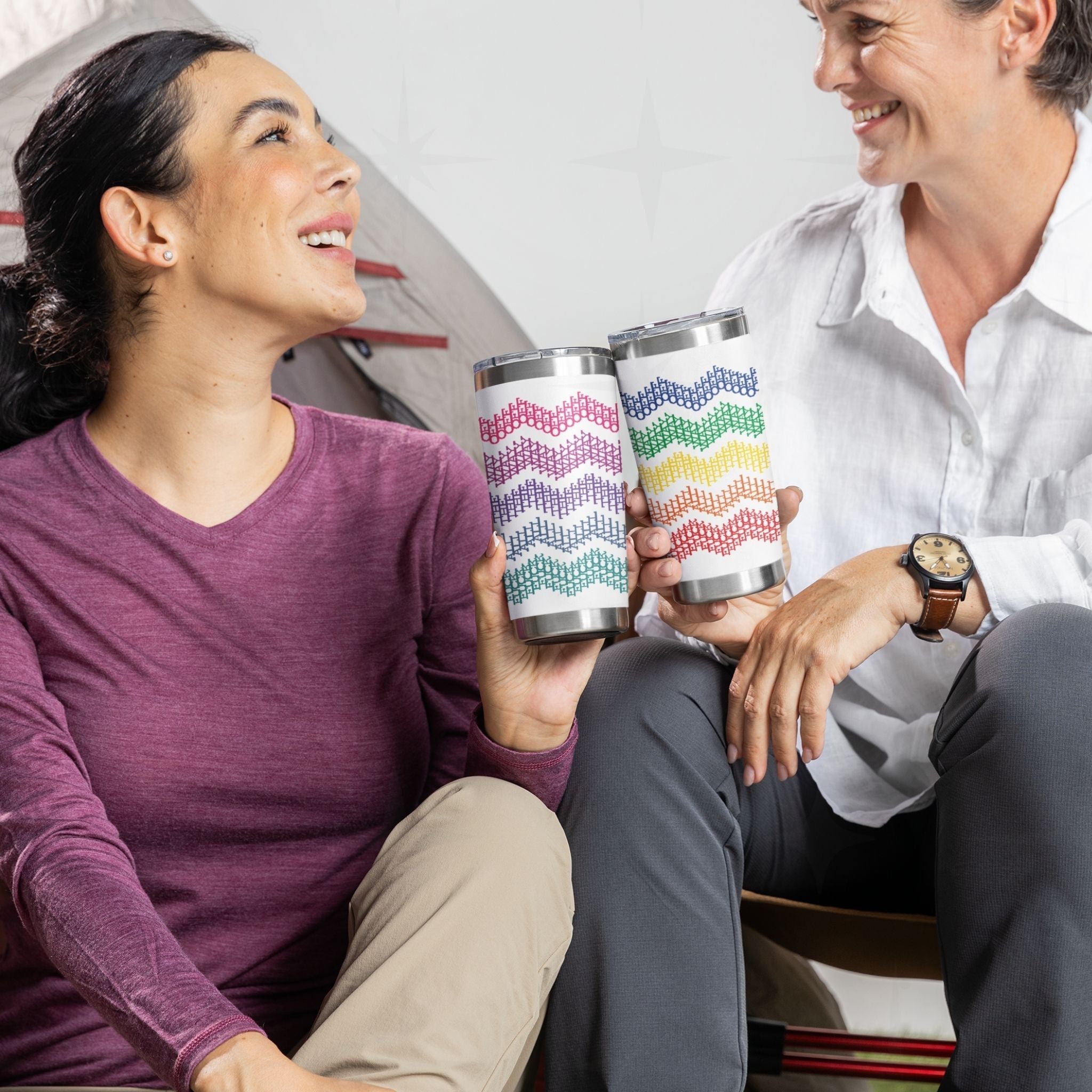 Two people sitting together holding bright stripe and rainbow stripe hidden profanity tumblers with cross stitch chevron pattern