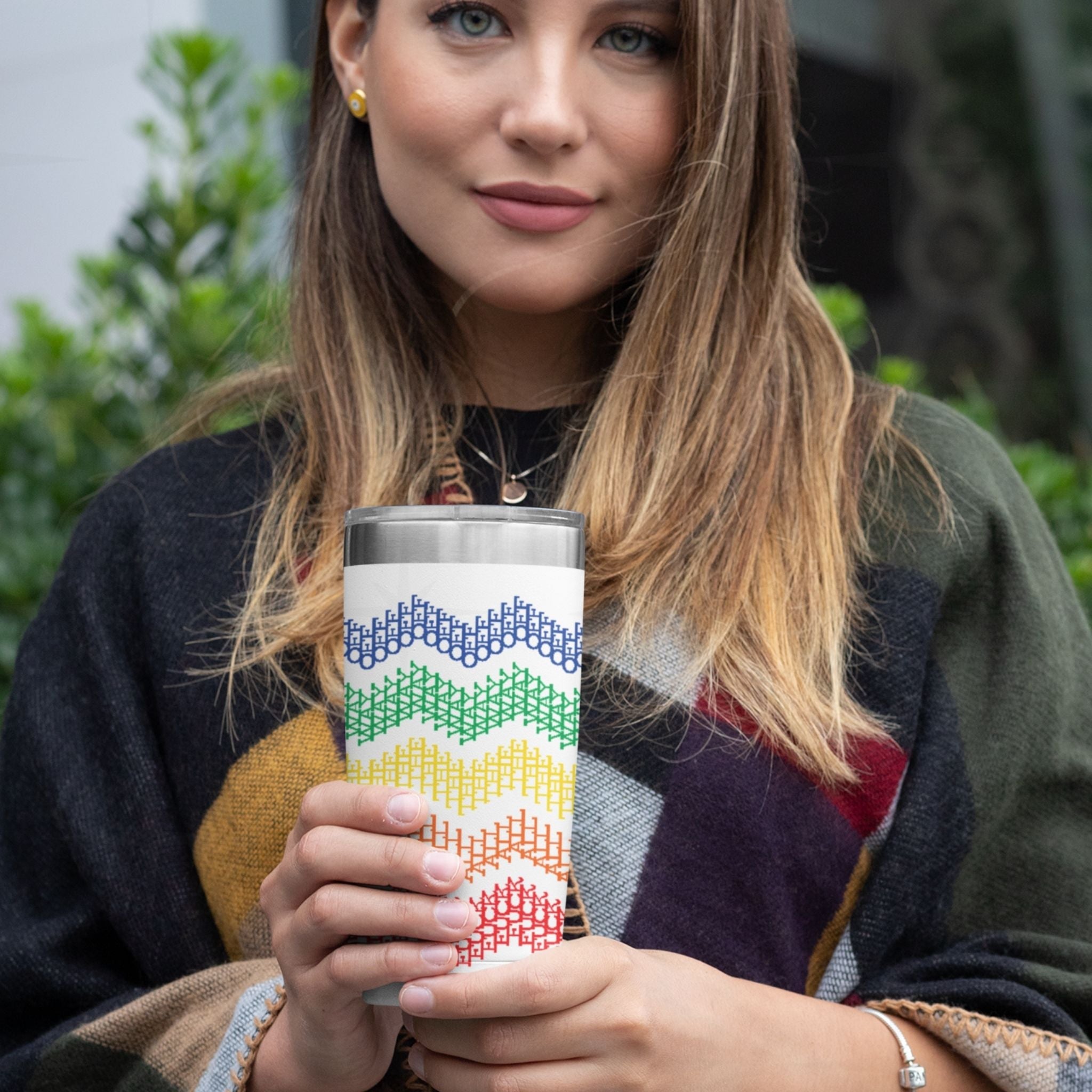 Person holding rainbow stripe hidden profanity tumbler outdoors near tree trunk showing cross stitch chevron pattern