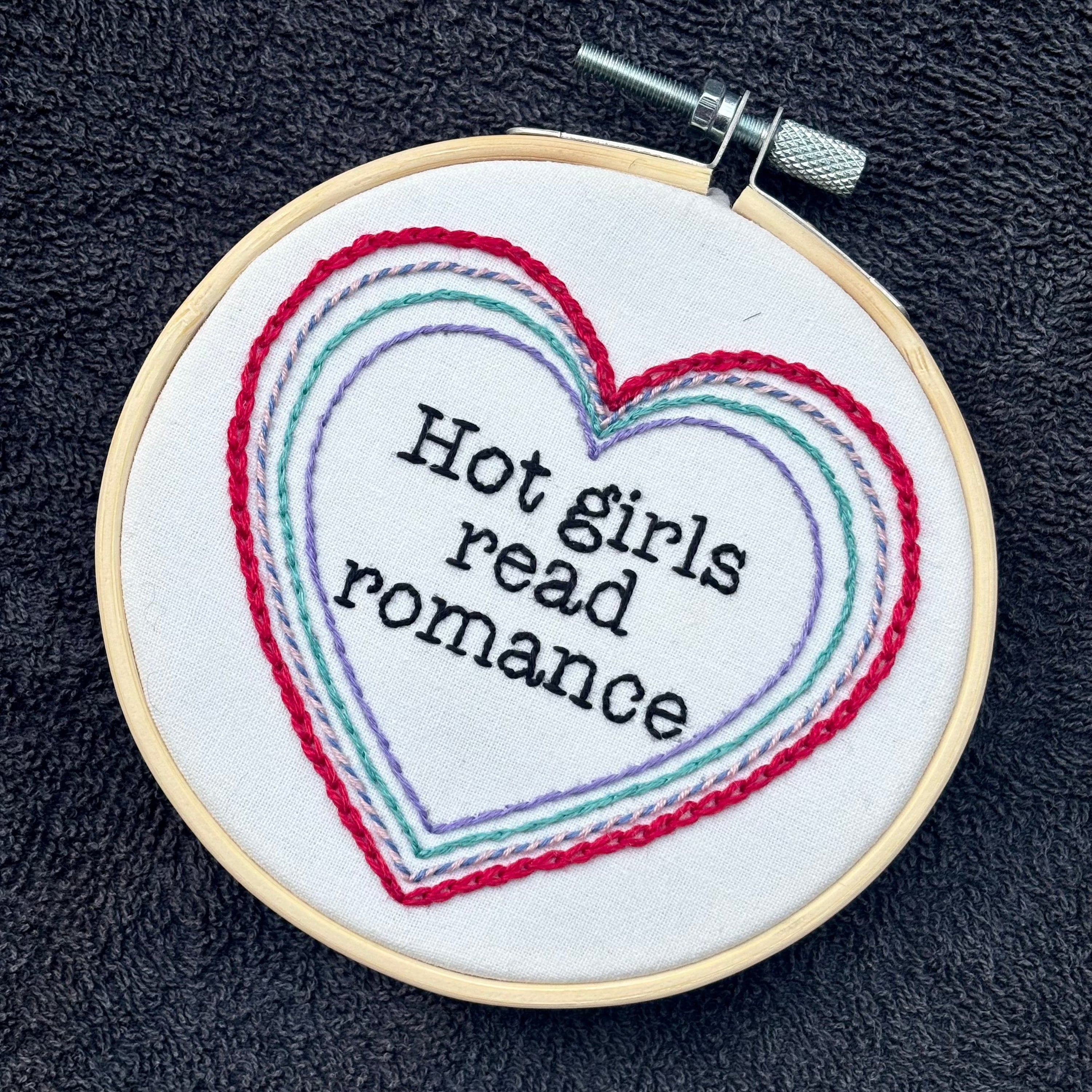 "Hot people read books" Quote hand embroidered in black embroidery thread in a serif, typewriter font. Text is framed in a series of hearts nested inside of each other and sewn in red, blue, pink, green, and purple. Each heart is stitched in a different hand embroidery stitch, as a sampler. Fabric is white cotton. Displayed as wall art in a light wooden embroidery hoop. Also sized for a christmas ornament.