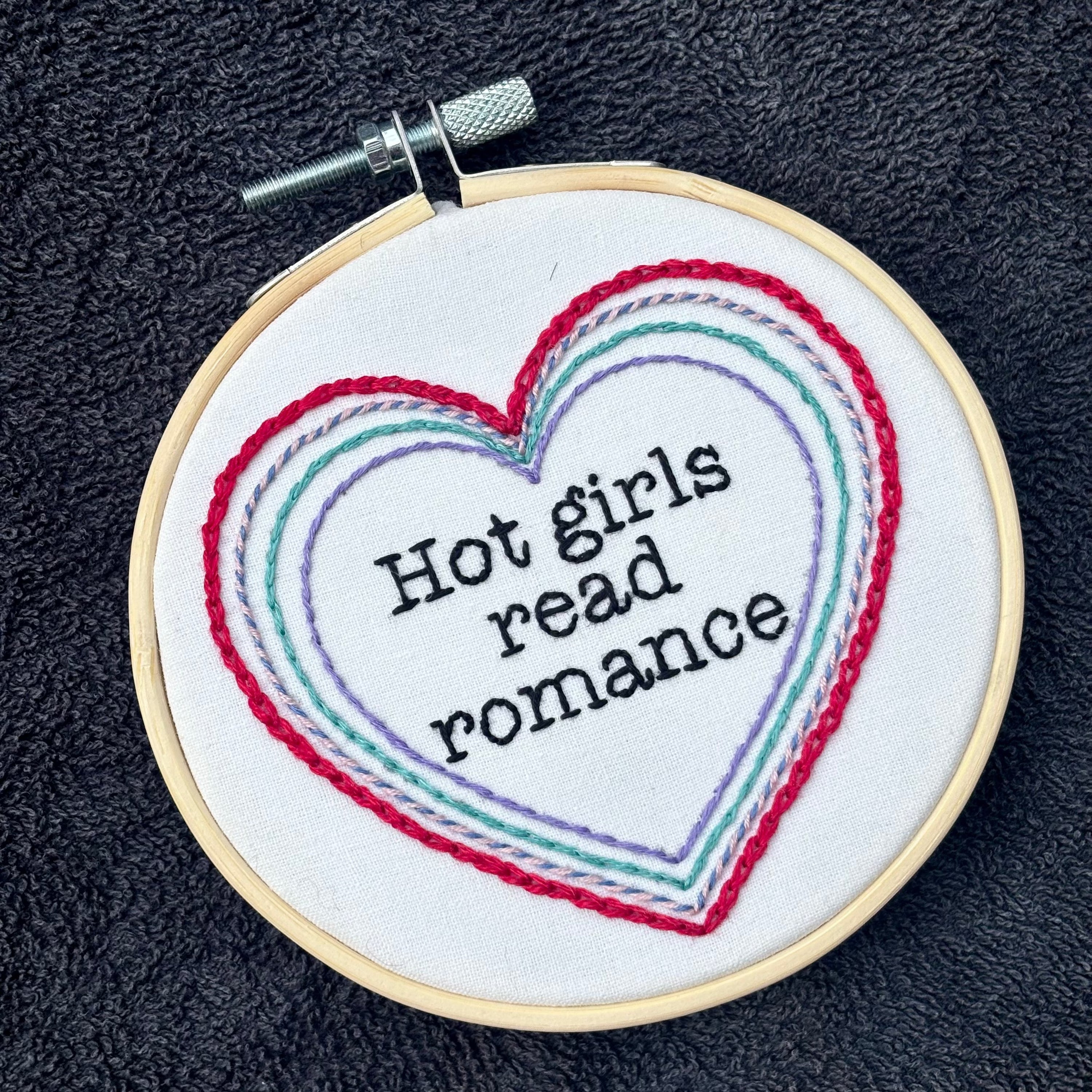 "Hot people read books" Quote hand embroidered in black embroidery thread in a serif, typewriter font. Text is framed in a series of hearts nested inside of each other and sewn in red, blue, pink, green, and purple. Each heart is stitched in a different hand embroidery stitch, as a sampler. Fabric is white cotton. Displayed as wall art in a light wooden embroidery hoop. Also sized for a christmas ornament.