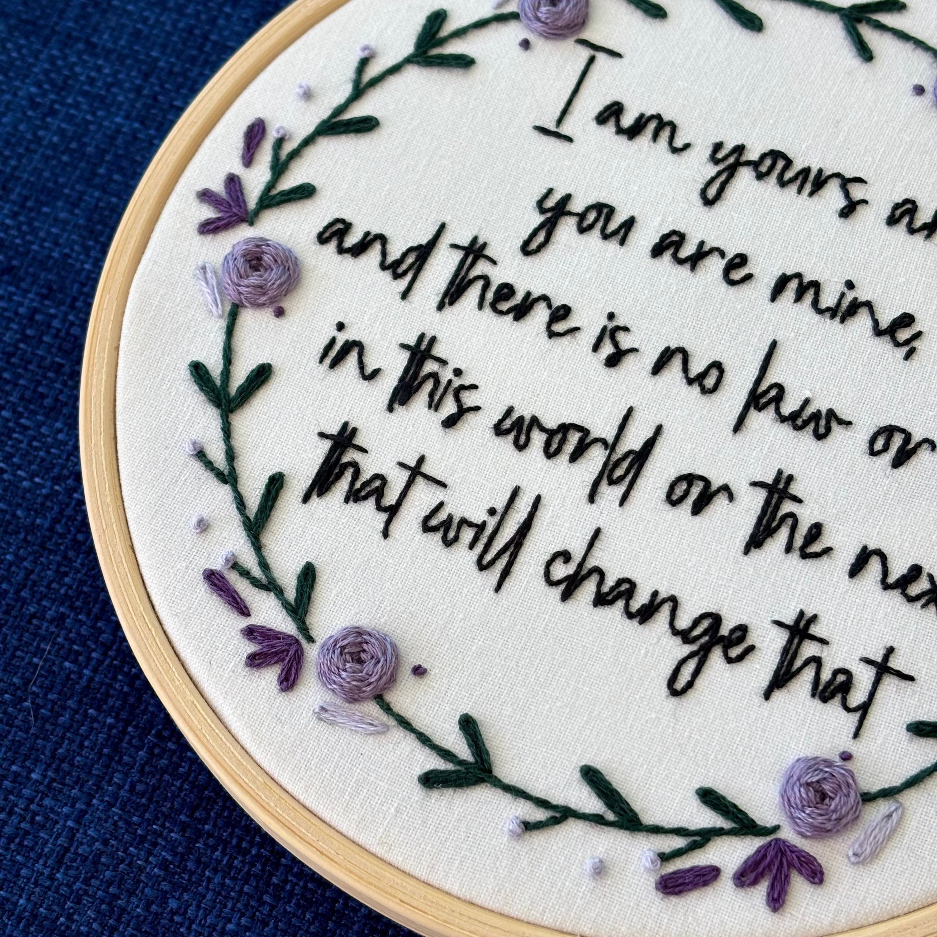 "I am yours and you are mine, and there is no law or rule in this world or the next that will change that." Quote hand embroidered in strong handwriting font in black embroidery floss. Surrounding the text is a wreath of dark green stems and leaves with lavender, violet, and deep purple flower petals, roses, and rose buds. Sewn on white cotton. Framed as wall art in a light wood embroidery hoop.