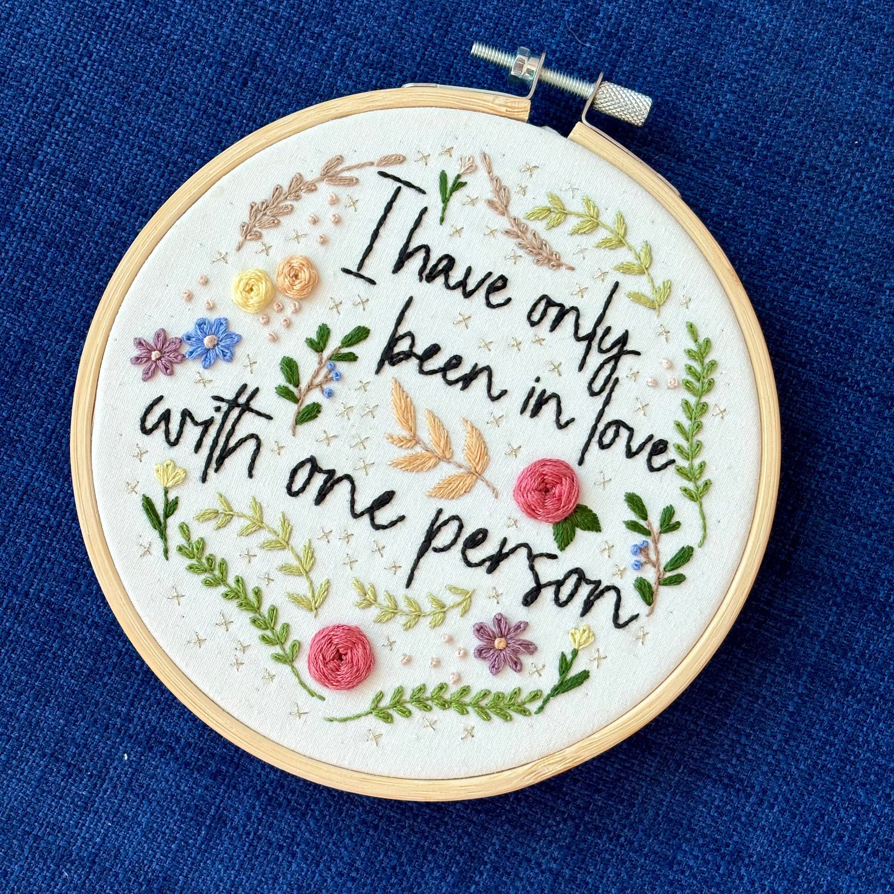 "I have only been in love with one person." Quote hand embroidered in handwriting font in black embroidery floss. Surrounding the text are vines and flowers and plants in shades of green, light browns, peach, pink, periwinkle blue, and dusky purple. Background is filled with pink dots and metallic gold Xs. Sewn on white cotton. Framed as wall art in a light wood embroidery hoop.