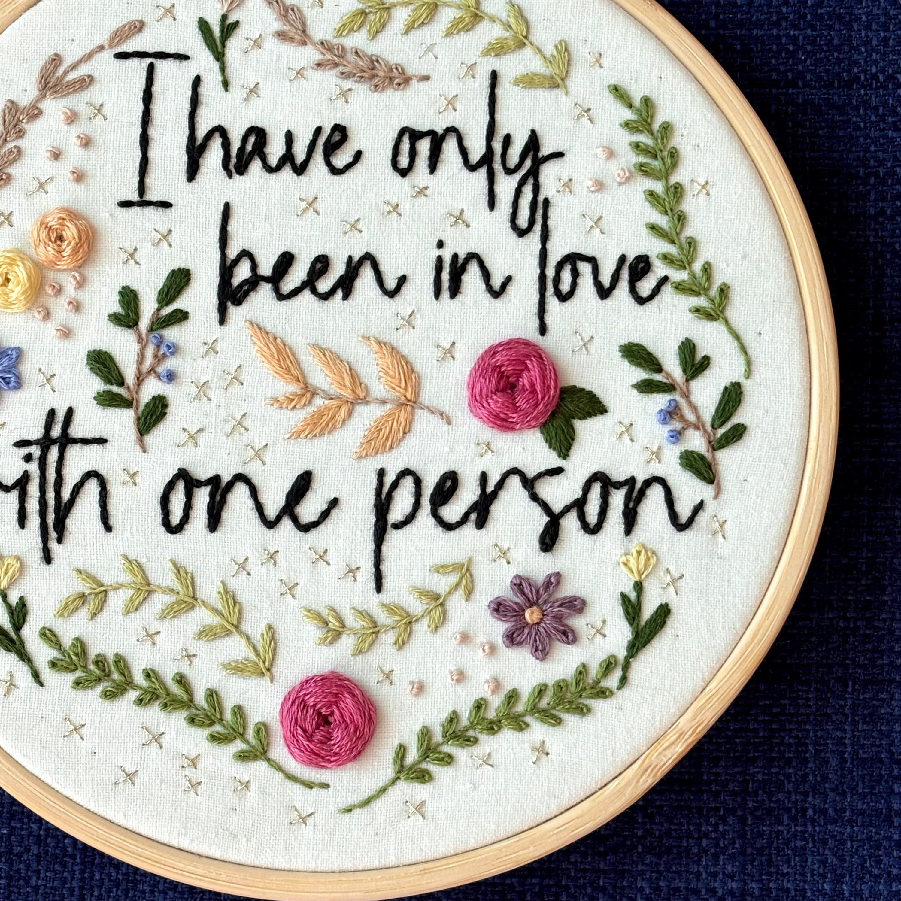 "I have only been in love with one person." Quote hand embroidered in handwriting font in black embroidery floss. Surrounding the text are vines and flowers and plants in shades of green, light browns, peach, pink, periwinkle blue, and dusky purple. Background is filled with pink dots and metallic gold Xs. Sewn on white cotton. Framed as wall art in a light wood embroidery hoop.