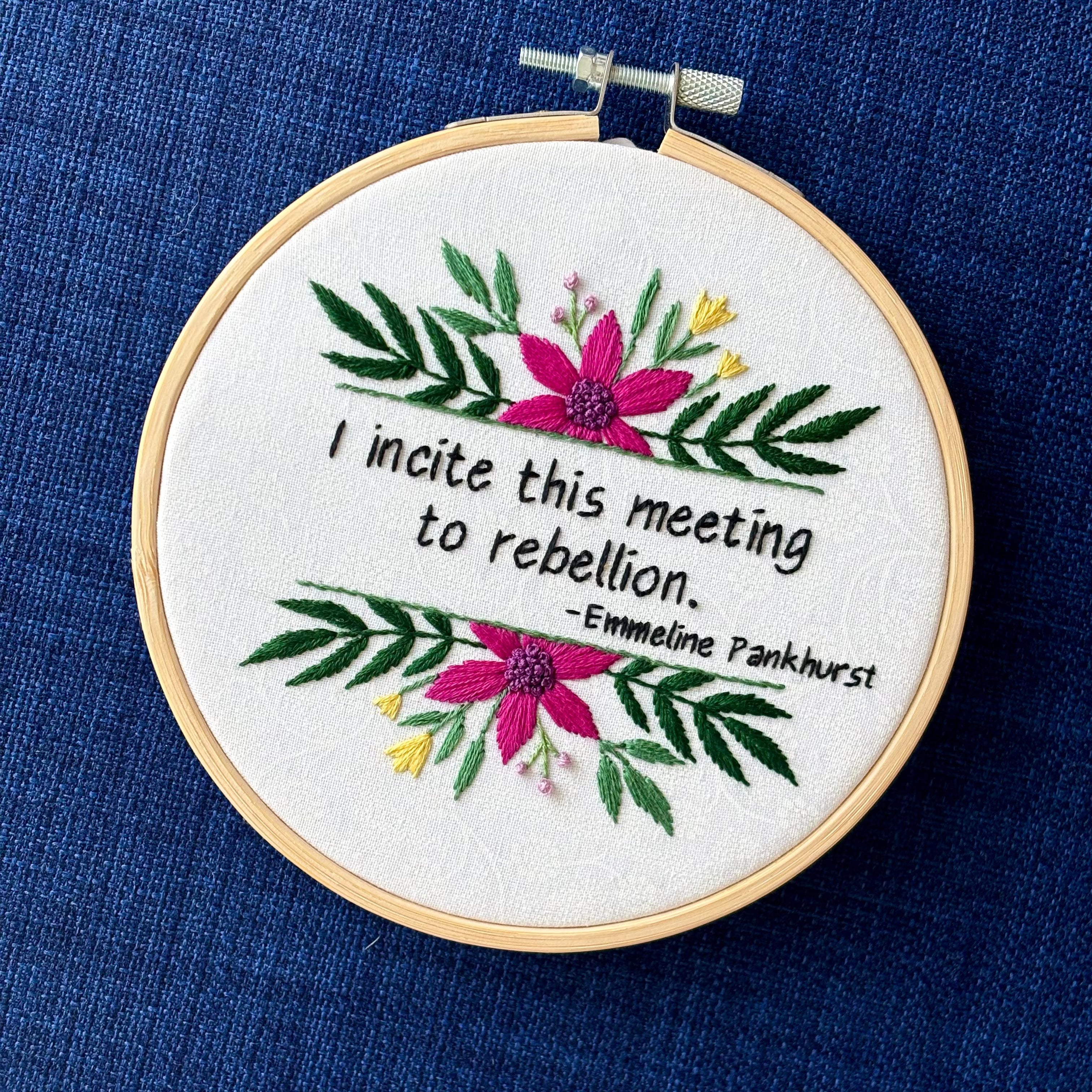 "I incite this meeting to rebellion - Emmeline Pankhurst". Quote hand embroidered in black thread. Framed with a fuchsia daisies, small yellow tulips, sprigs of green leaves, and branches with lavender flower buds. Hand sewn on white-on-white cotton and displayed as wall art in a light wooden embroidery hoop.