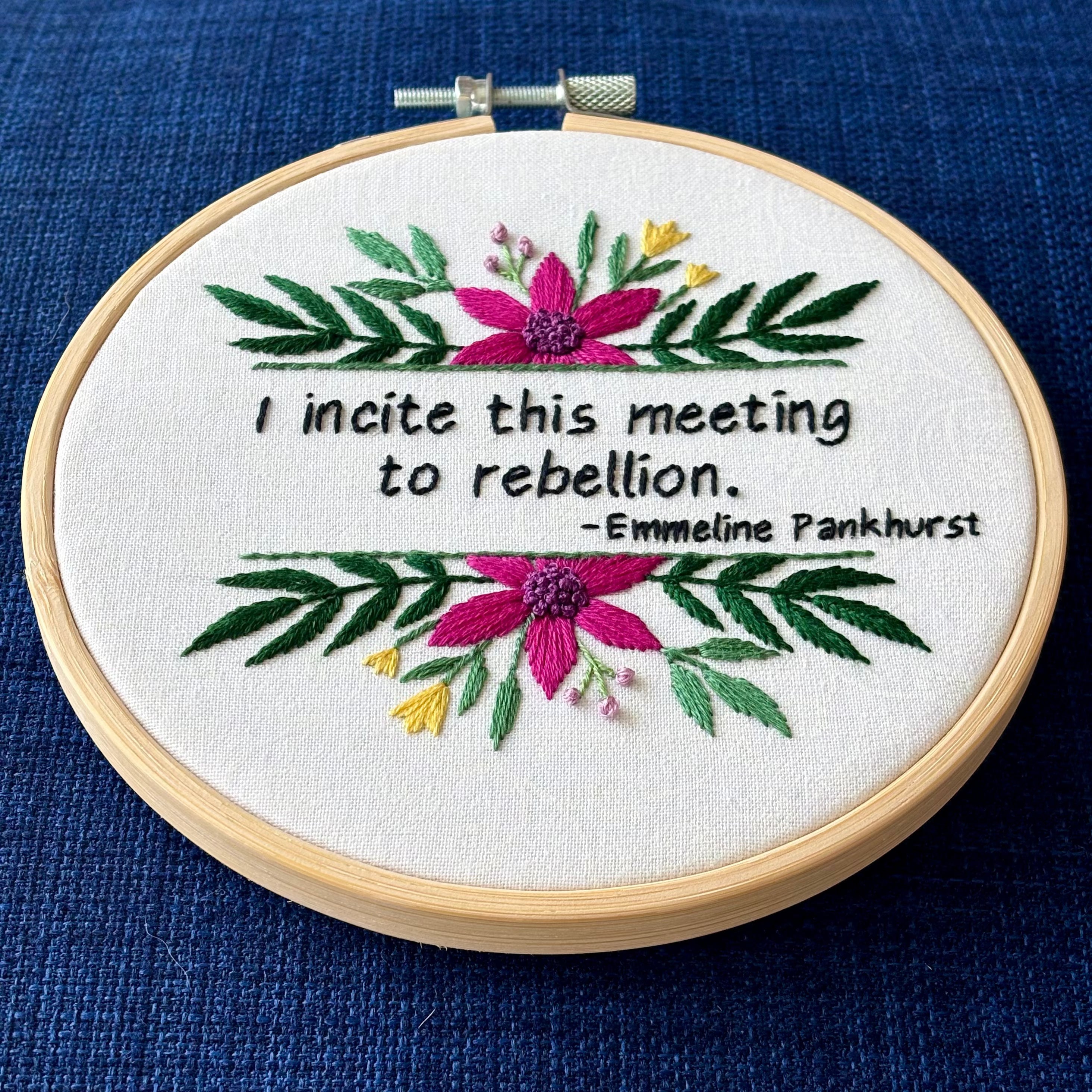 "I incite this meeting to rebellion - Emmeline Pankhurst". Quote hand embroidered in black thread. Framed with a fuchsia daisies, small yellow tulips, sprigs of green leaves, and branches with lavender flower buds. Hand sewn on white-on-white cotton and displayed as wall art in a light wooden embroidery hoop.