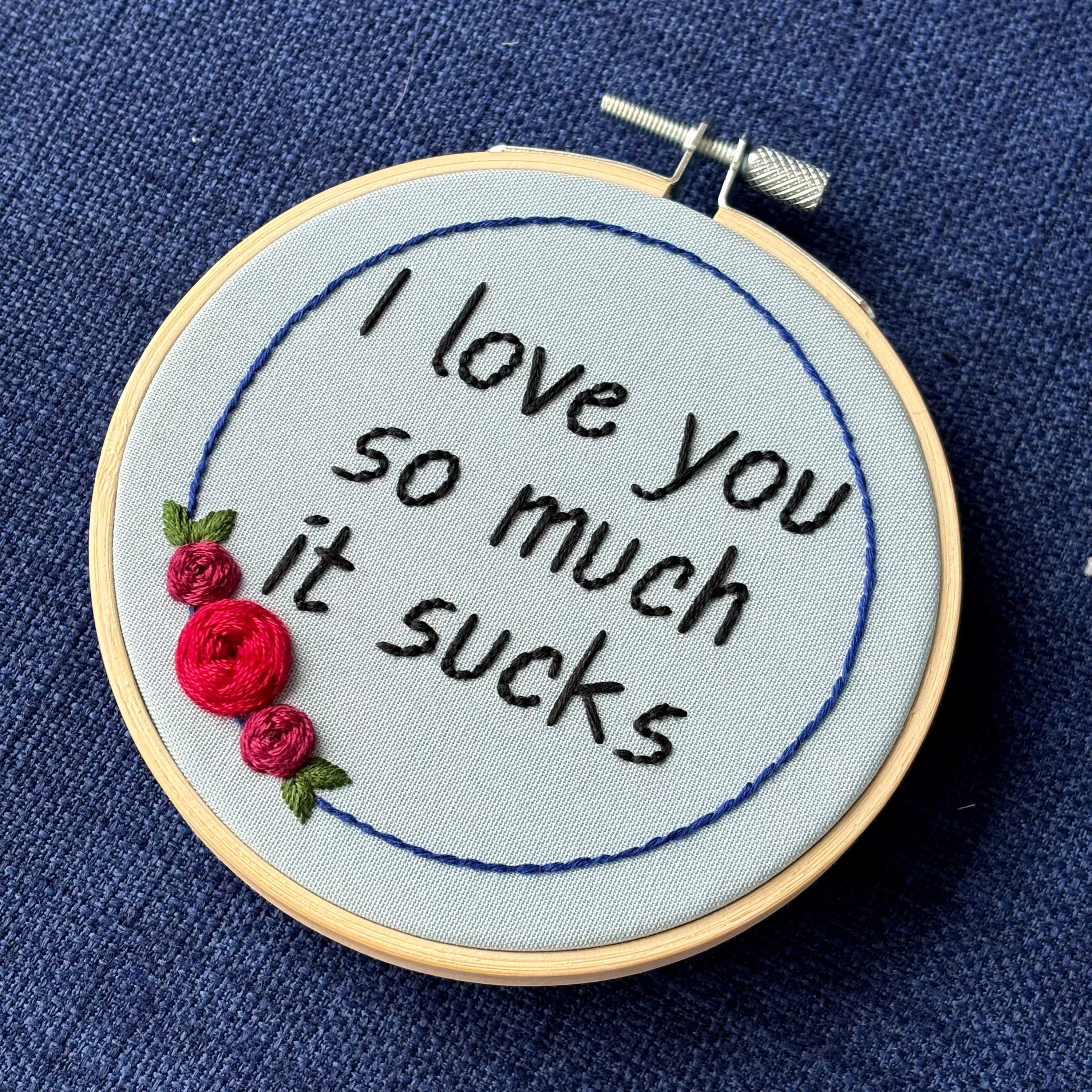 "I love you so much it sucks." Quote hand embroidered in handwriting font in black embroidery thread. Around the text is a floral wreath of a blue ring, red roses and green leaves. Fabric is light blue, and completed embroidery is displayed in a light wood embroidery hoop.