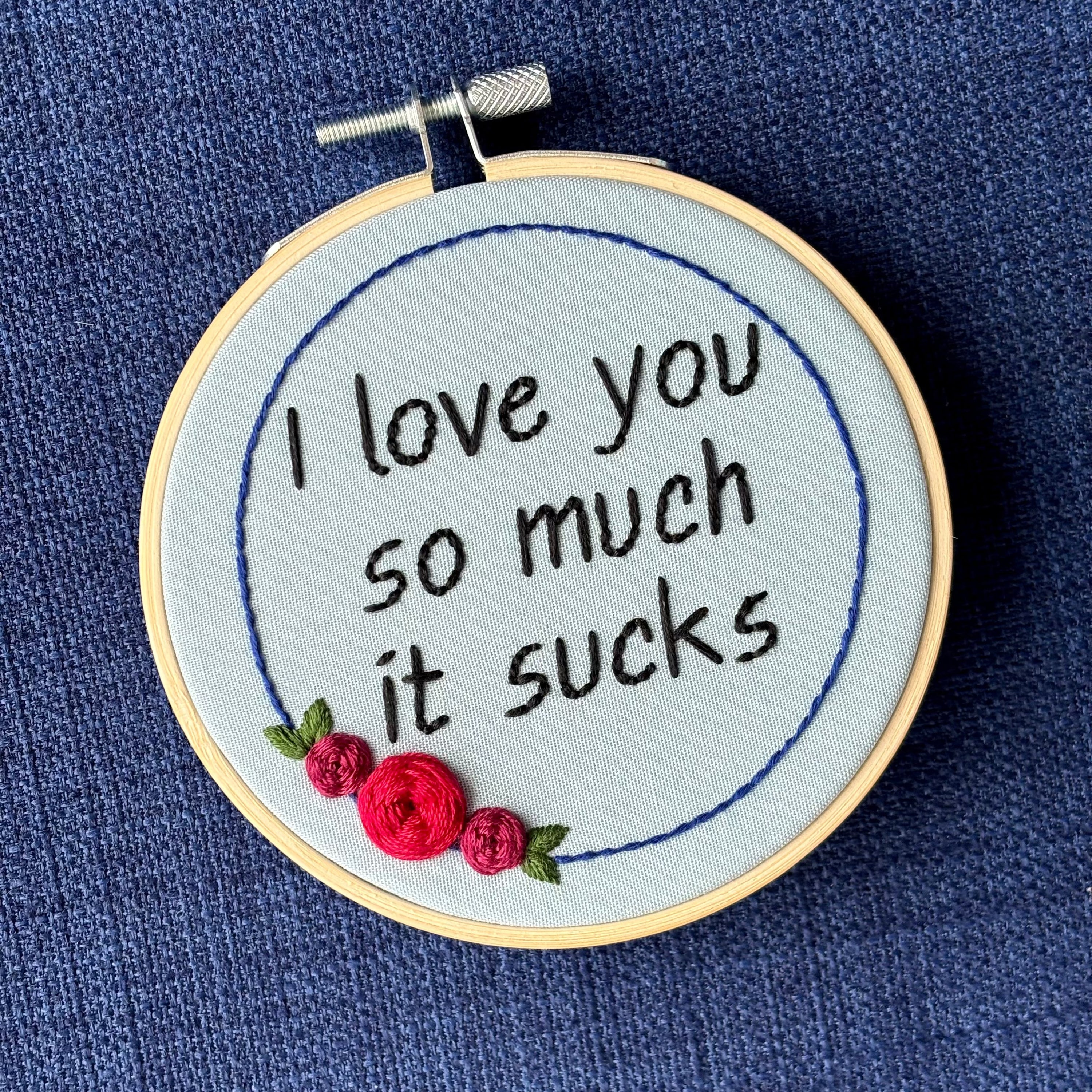 "I love you so much it sucks." Quote hand embroidered in handwriting font in black embroidery thread. Around the text is a floral wreath of a blue ring, red roses and green leaves. Fabric is light blue, and completed embroidery is displayed in a light wood embroidery hoop.