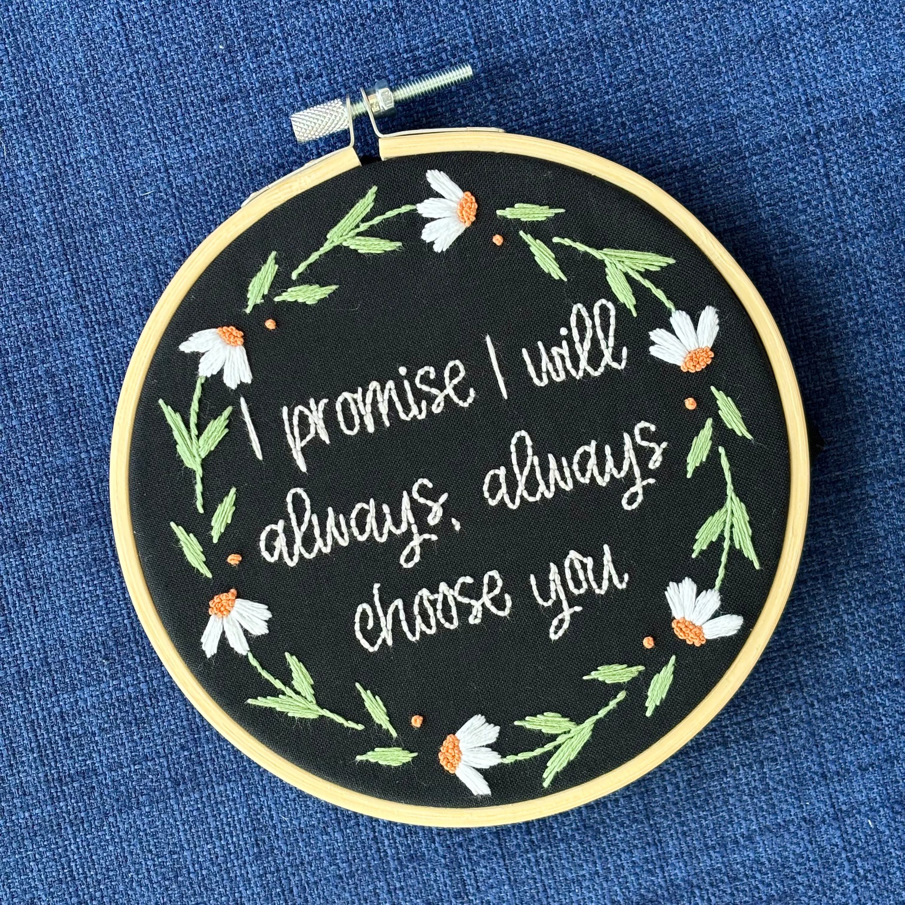 "I promise I will always, always choose you." Quote hand embroidered in handwriting font in the palest blush pink. Surrounding the text is a floral wreath of white daisies with orange centers. Sewn on black cotton. Framed in a light wood embroidery hoop as wall art.