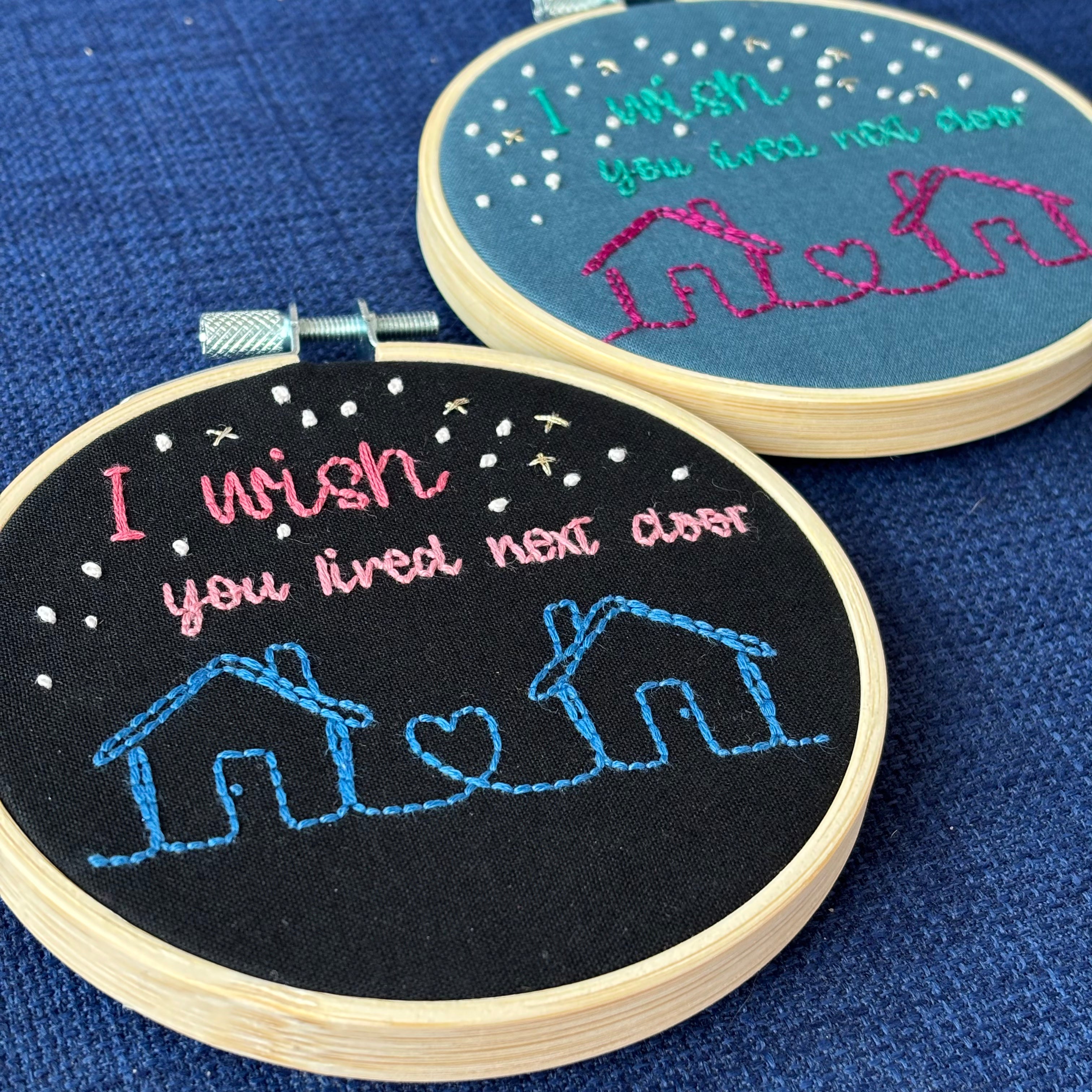 2 Christmas ornaments framed in light wooden embroidery hoops. One ornament has two line drawing houses connected with a heart, hand stitched in pink thread. The sky has white and gold stars. Teal text reads, "I wish you lived next door." Cotton fabric is blue. The second handmade Christmas ornament is the same design but the 2 houses connected with a heart are hand stitched in blue thread. The sky has white and gold stars. Pink text reads, "I wish you lived next door." Fabric is black cotton.