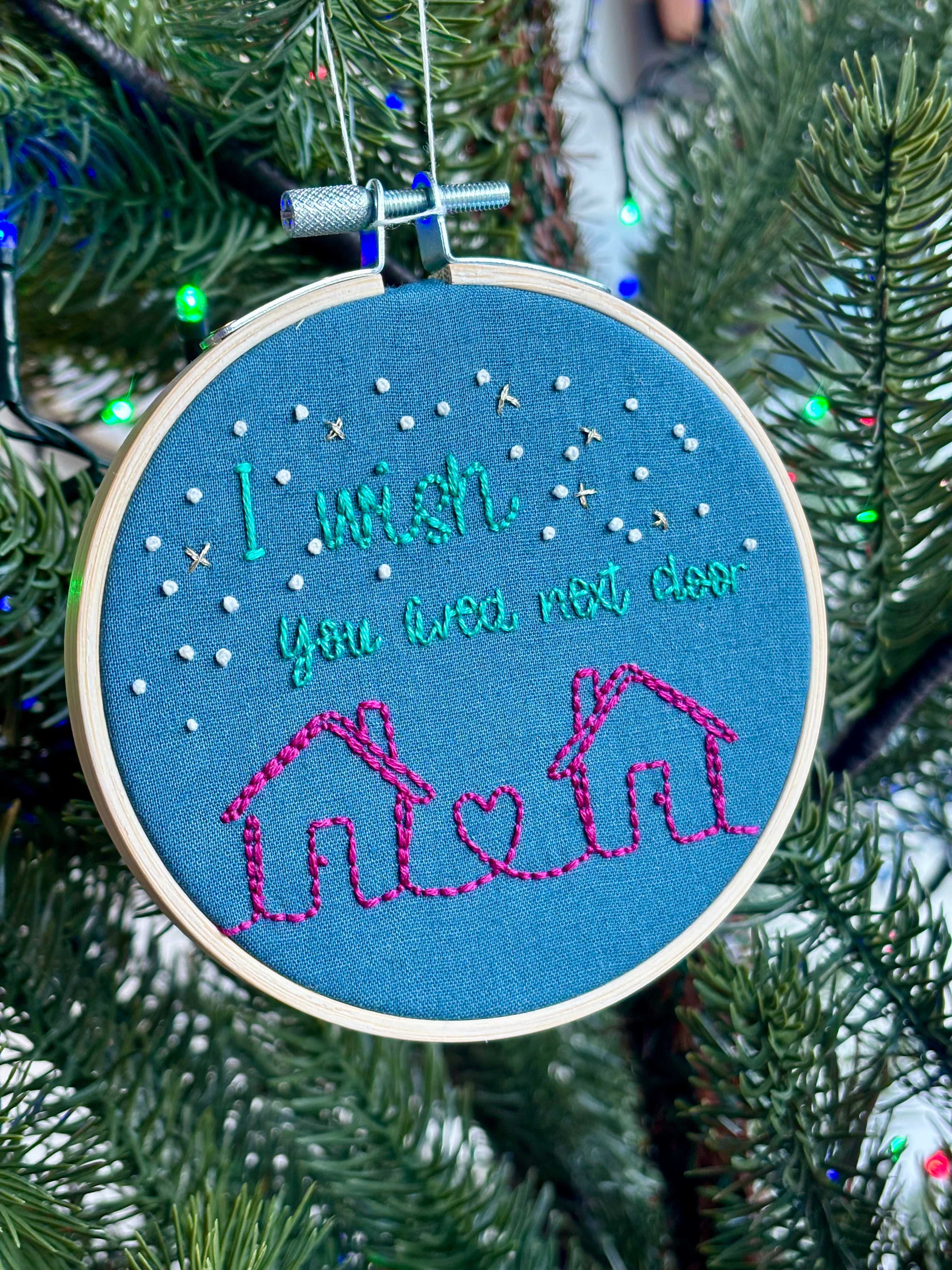 PDF Pattern. Two line drawing houses connected with a heart, hand stitched in bright pink thread. The sky has white and gold stars. Teal text reads, "I wish you lived next door." Cotton fabric is medium blue. Finished Christmas ornament is framed in a light wooden embroidery hoop and hung on a Christmas tree.