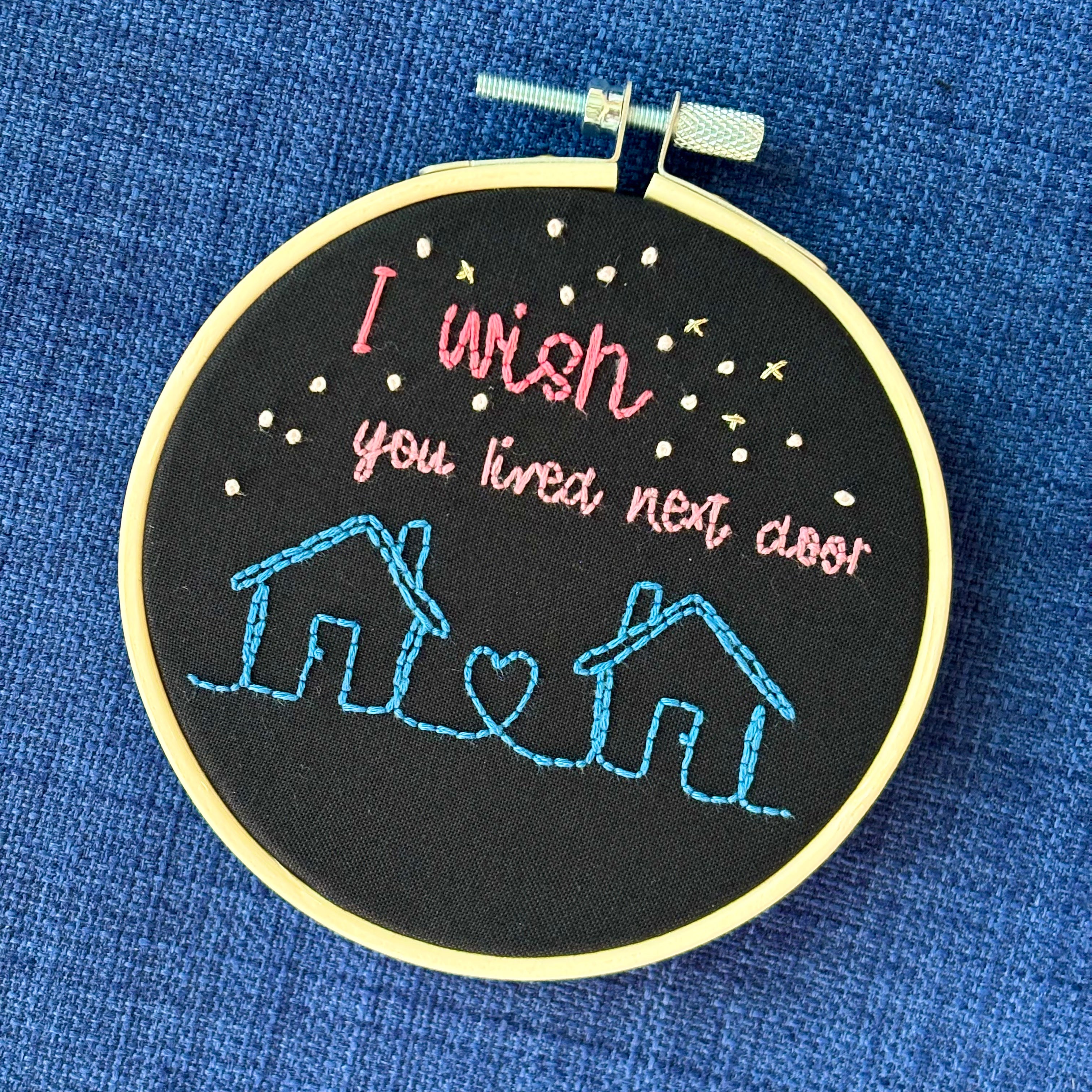 Two line drawing houses connected with a heart, hand stitched in blue thread. The sky has white and gold stars. Pink text reads, "I wish you lived next door." Fabric is black cotton. Finished Christmas ornament is framed in a light wooden embroidery hoop.