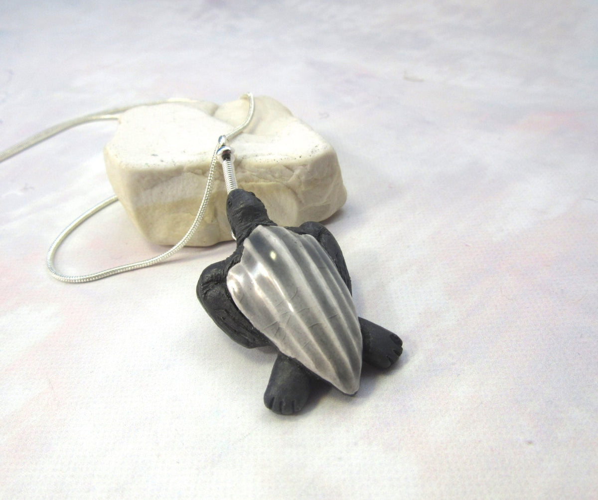 The Hand Painted Leatherback Turtle Pendant: Ocean Jewelry features a metallic silver shell with black accents, displayed on a light stone—a striking, ocean-inspired necklace set against a pale background.