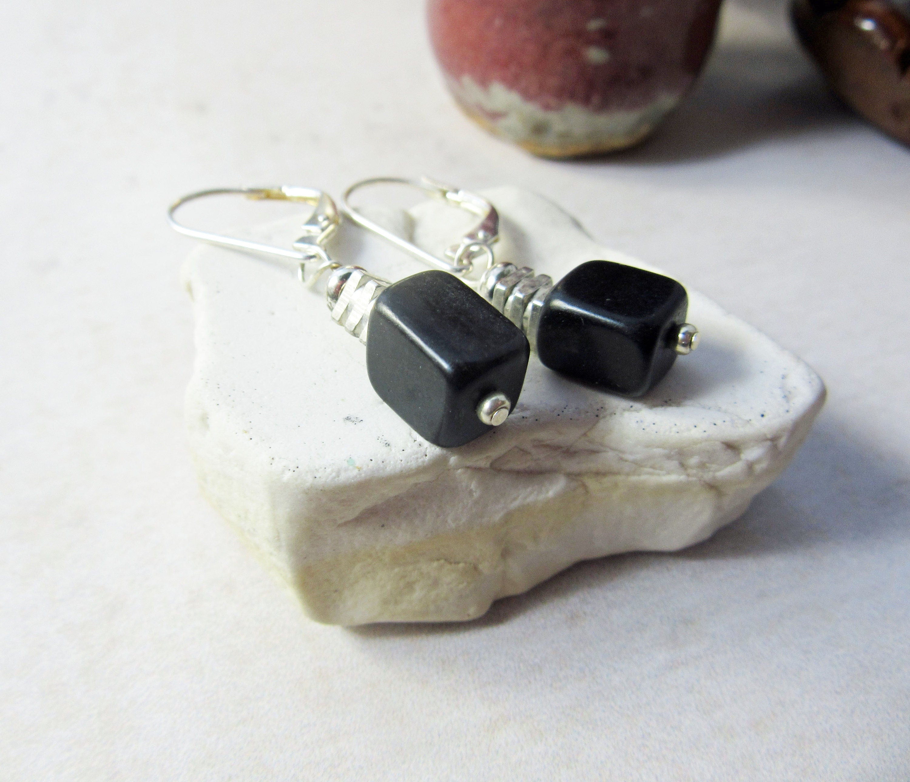 Black Stone Drop Earrings: Minimalist Silver Cube Jewelry rest on a white textured stone, showcasing their simple silver hooks and square black bead accents, with a blurred ceramic object in the background.