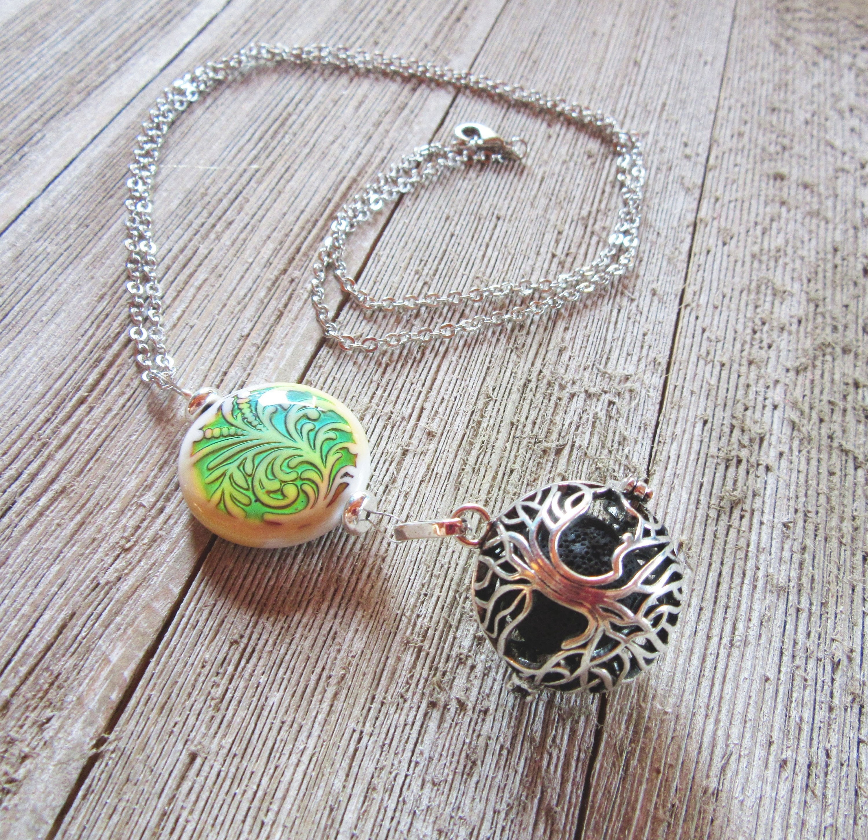 The Color Changing Tree of Life Aromatherapy Necklace is a handmade silver essential oil diffuser with a round yellow pendant, green leaf accent, and filigree sphere, elegantly displayed on wood.