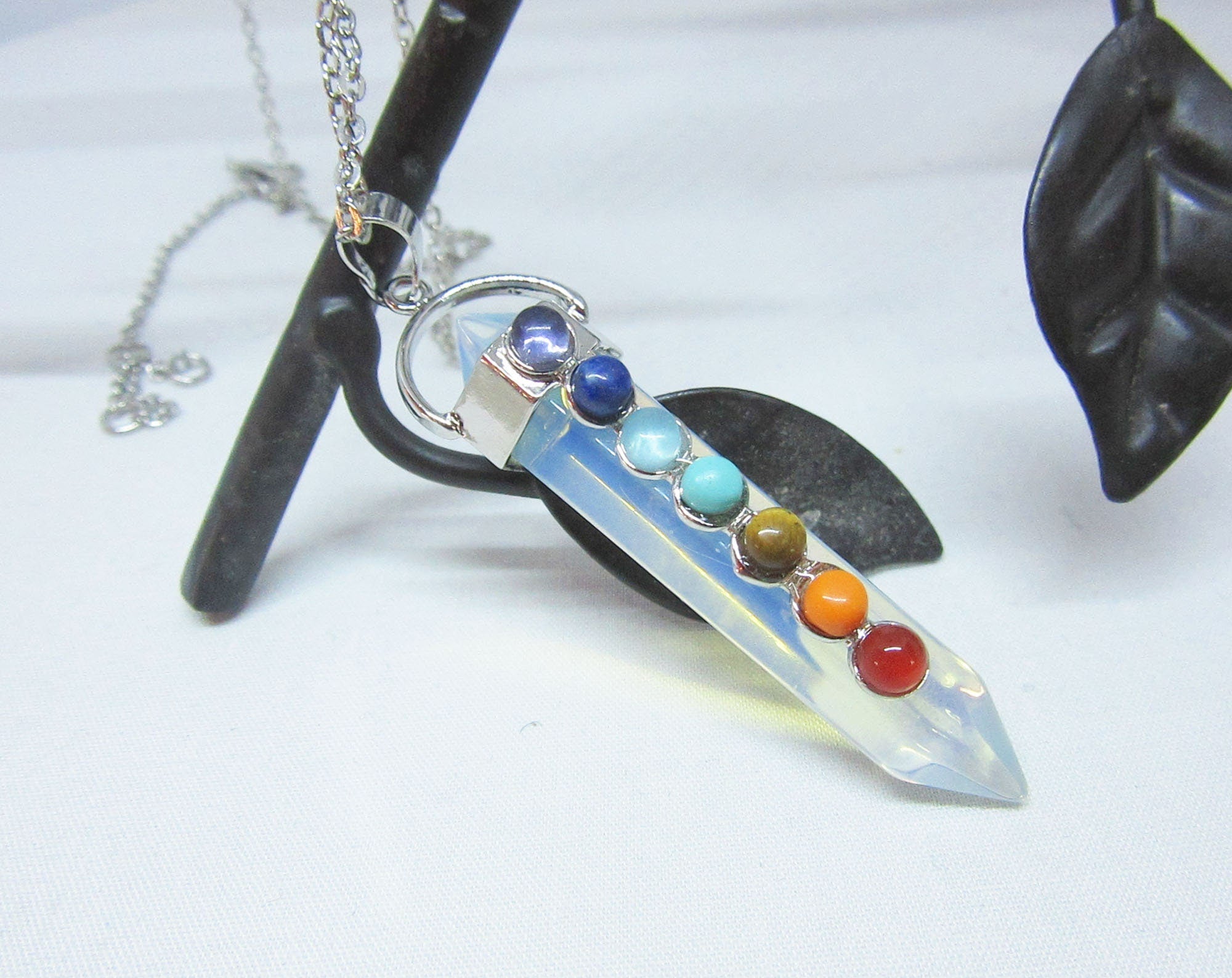 The Handcrafted Opalite Wand Pendant: Chakra Stone Necklace features seven colorful chakra stones on a silver chain, elegantly displayed over a black metal leaf—a distinctive piece of handmade jewelry.