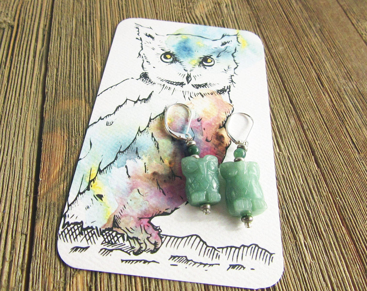 Handcrafted Owl Earrings: Natural Green Stone, Sterling Silver-Filled, are displayed on a card with a watercolor and ink owl illustration, all set atop a rustic wooden surface.