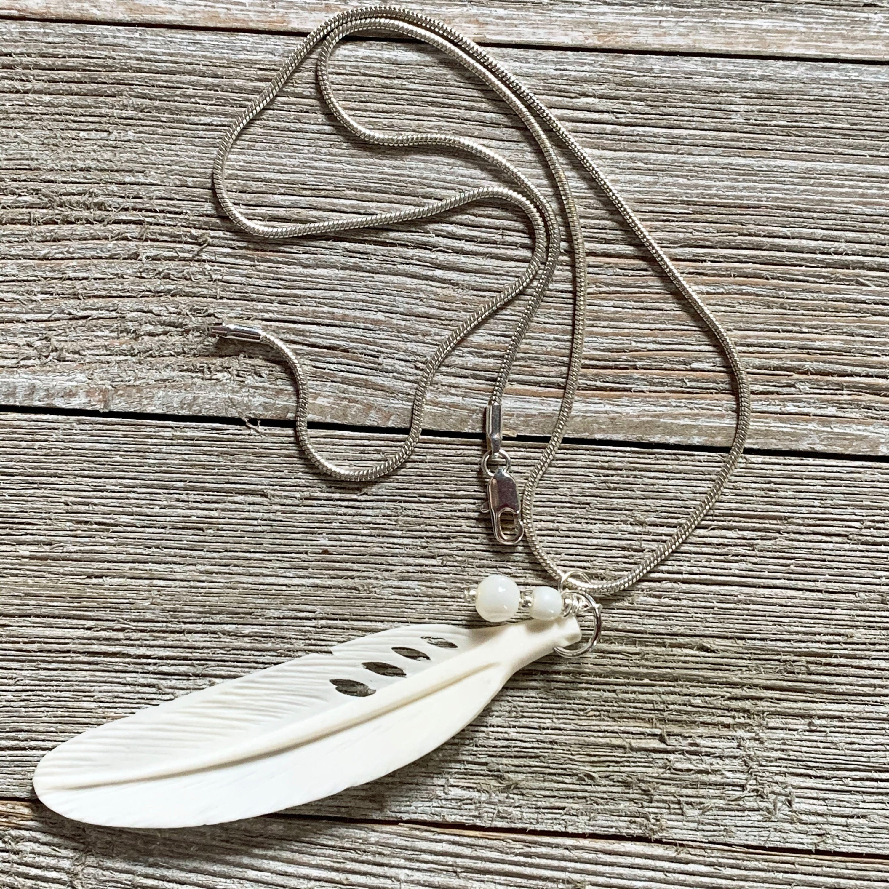 The White Feather Necklace features an angel sign carved bone and mother of pearl pendant, displayed on a rustic wooden surface—ideal for those seeking meaningful spiritual jewelry.