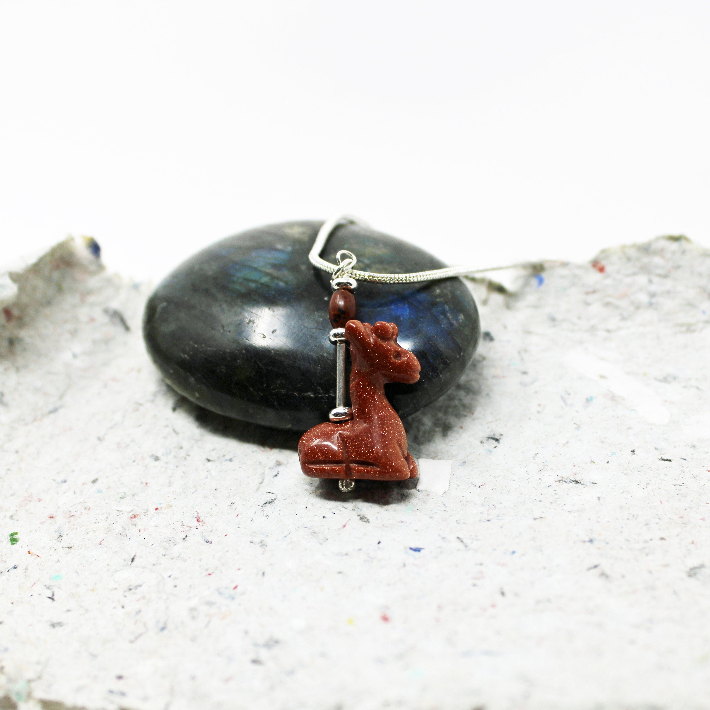 The Handcrafted Goldstone Giraffe Charm Pendant, designed for animal lovers, features a silver giraffe charm and goldstone pendant displayed on a textured gray surface with a polished dark stone in the background.