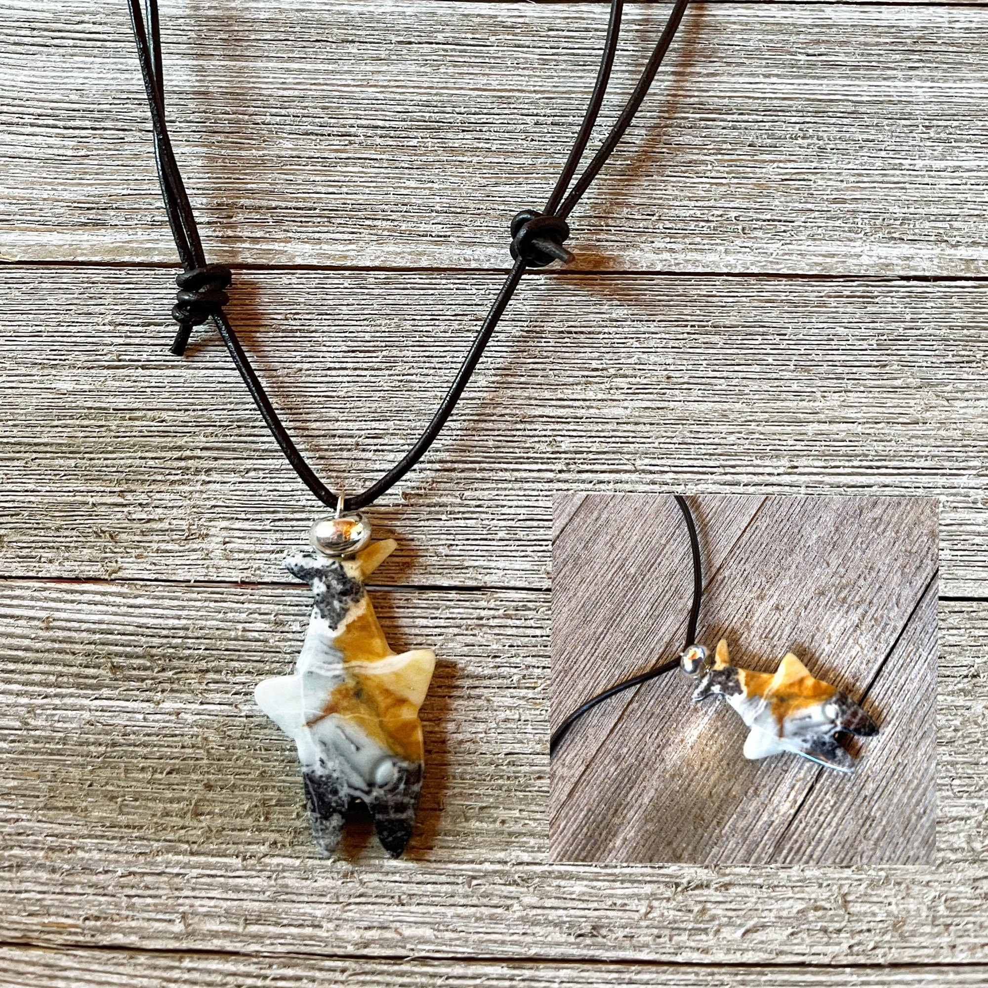 Displayed on a wooden surface, the Shark Pendant Necklace features a handcrafted amazonite shark-shaped pendant in black, white, and tan on a black cord. An inset offers a close-up of this unique ocean theme jewelry piece.