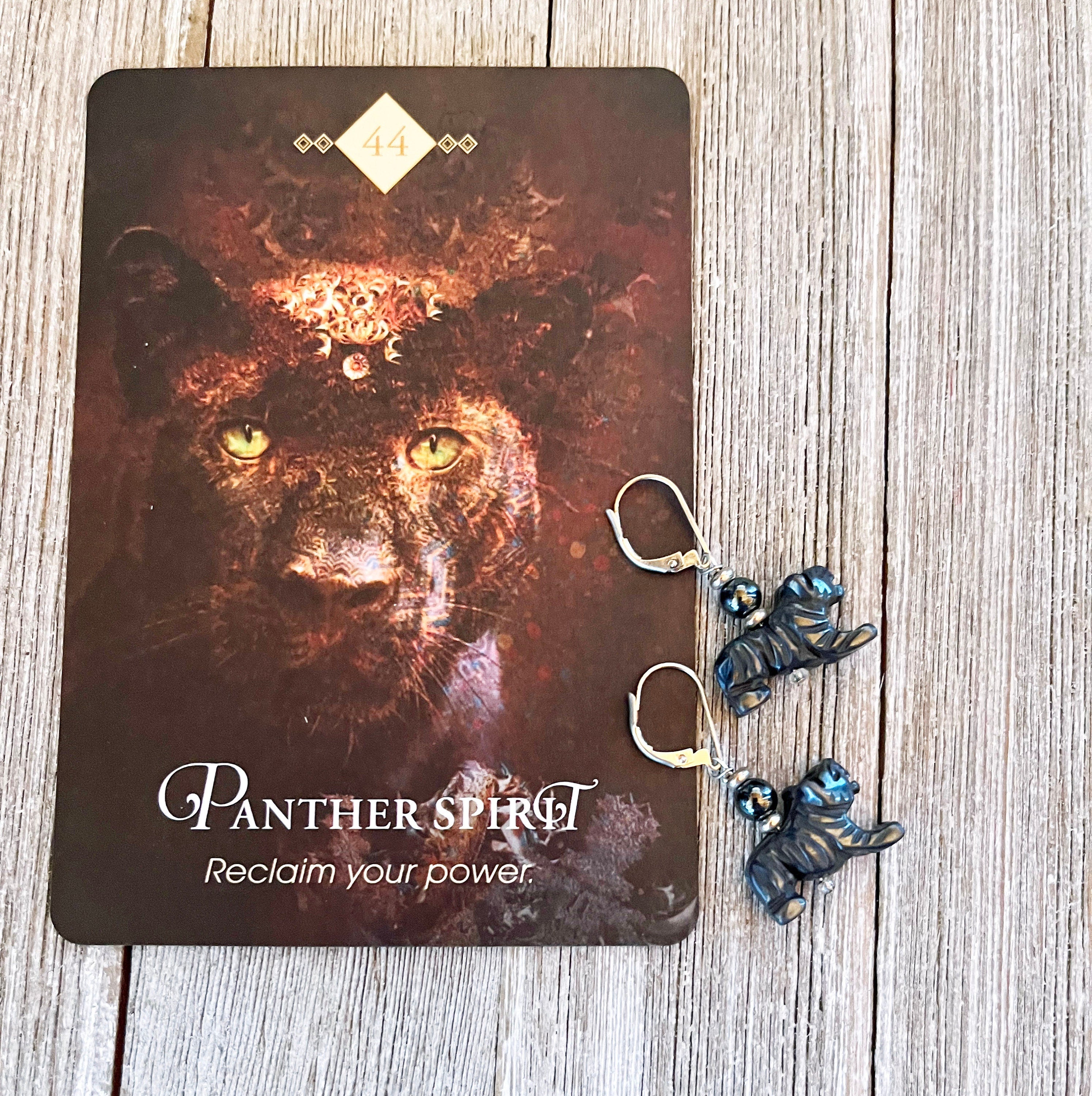 A card showing a black panther, "Panther Spirit," "Reclaim your power," and the number 44 rests on wood beside Enchanting Panther Hematite Earrings—unique animal jewelry perfect for big cat lovers with grounding blue accents.