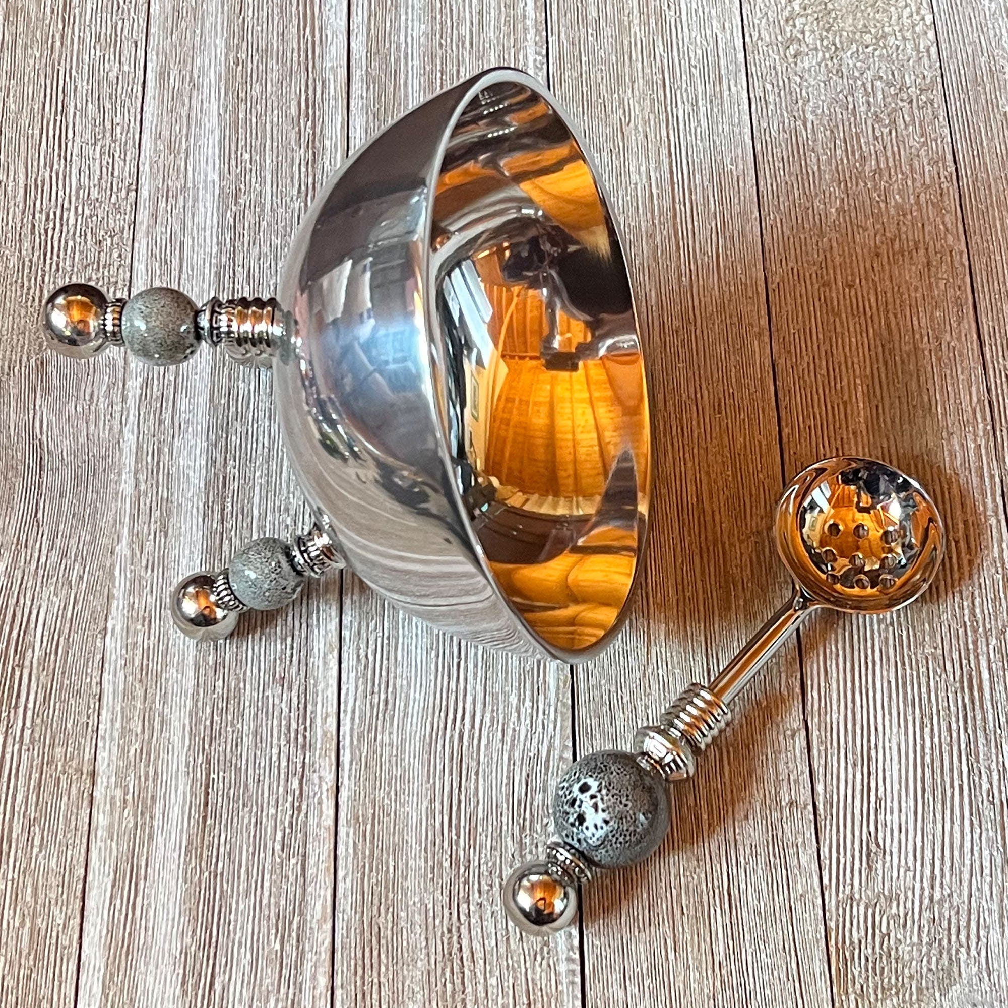 The Stainless Steel Bowl Set features gray ceramic legs and a matching spoon, both with intricate handle designs and reflective surfaces. Ideal as an altar bowl set or spiritual jewelry organizer, they rest elegantly on any wooden surface.