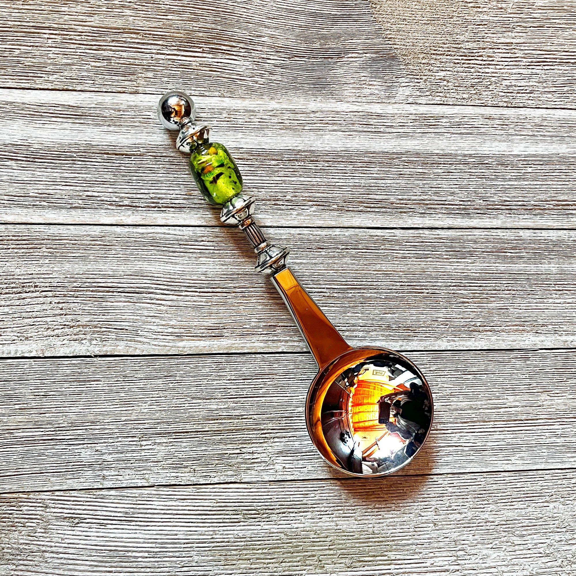 The Art Glass Beaded Handle Coffee Spoon, a stainless steel 1 Tbsp measure with a green art glass bead on its handle, rests on a textured wooden surface. The spoon’s bowl reflects colorful indoor lights and surroundings.