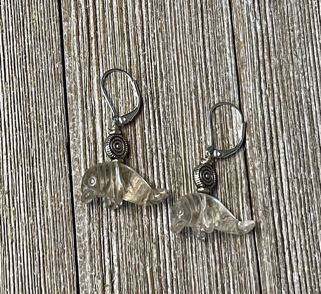 Manatee Earrings: Carved clear quartz charms with a decorative spiral hang from sterling silver lever backs, displayed on a wooden surface.