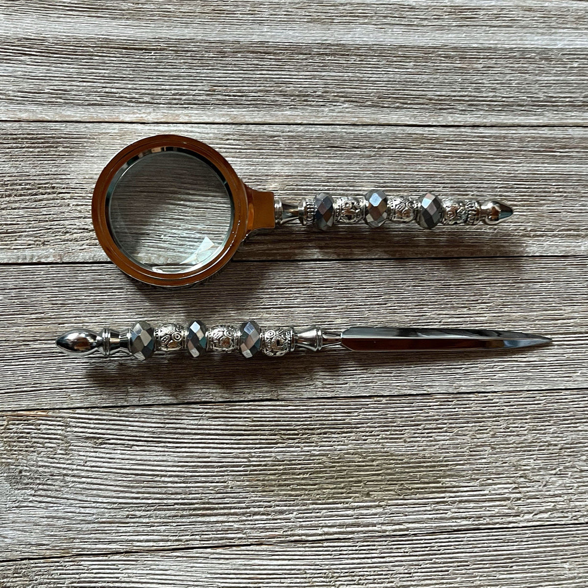 The Handmade Silver Desk Set features a magnifying glass and letter opener with faceted bead handles, shown here lying on a wooden surface.
