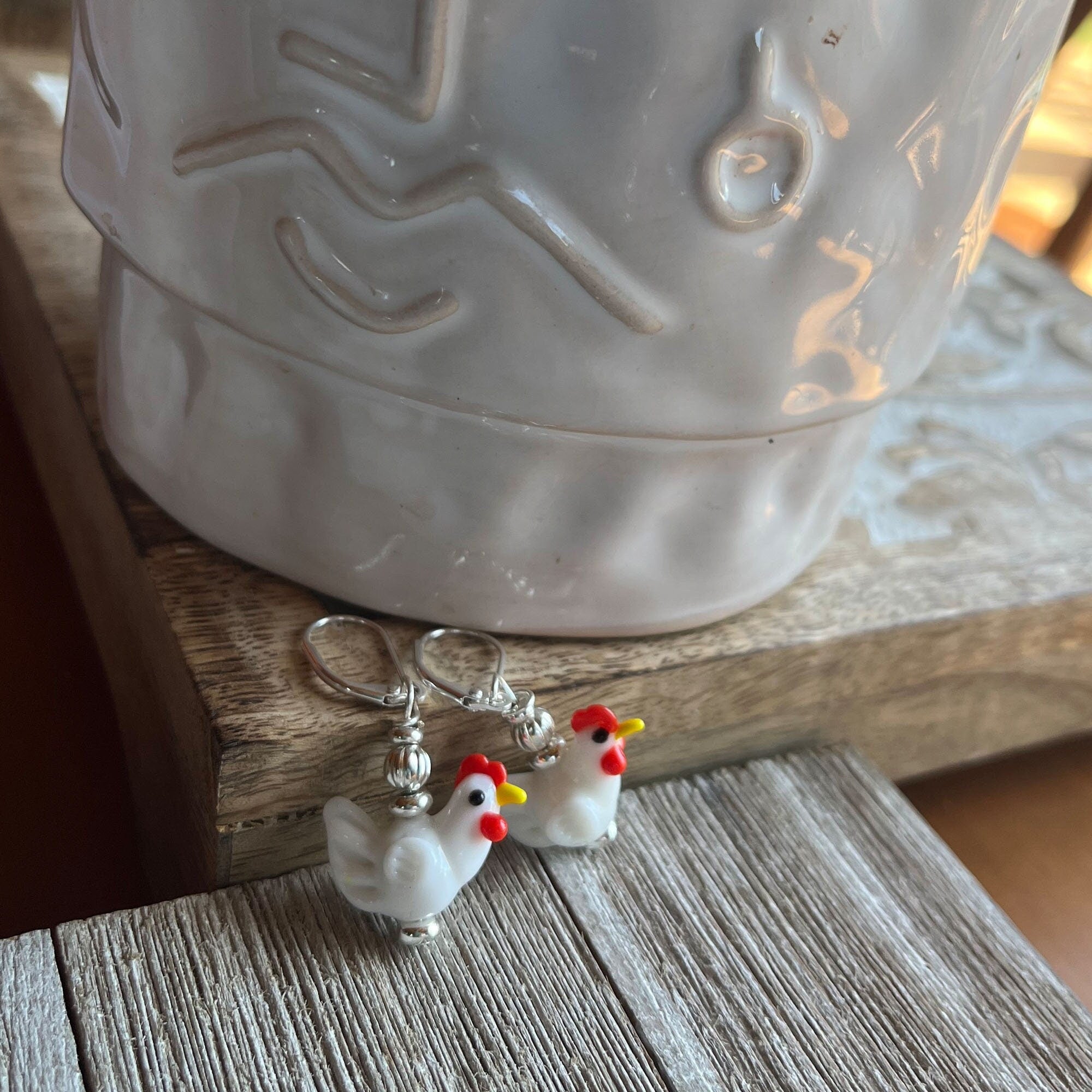 The Farm Charm Hen Earrings are handmade glass chicken jewelry featuring white hens with red combs and yellow beaks—the perfect gift for farm lovers. Displayed on a wooden surface in front of a ceramic pot.