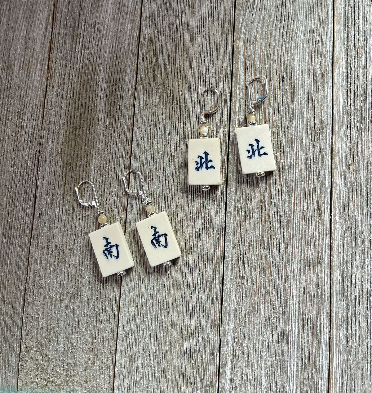 The Mahjong Tile Earrings: North/South Winds, perfect for mahjong players, feature four rectangular tiles with metallic hooks and black Chinese characters, arranged on a weathered wooden surface for a unique, Asian-inspired look.