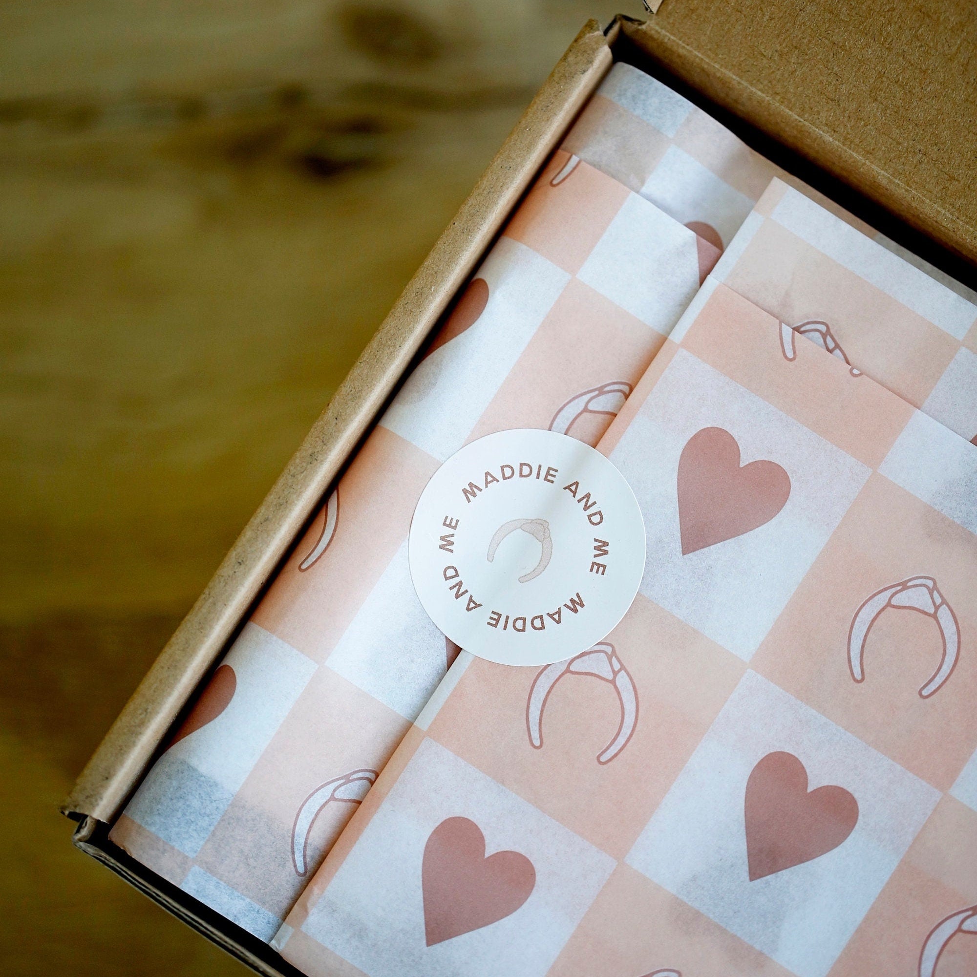 May include: A brown cardboard box with tissue paper featuring a pink and white checkered pattern with heart and headband illustrations. A round sticker with the text "MADDIE AND ME" and a headband illustration is on top.