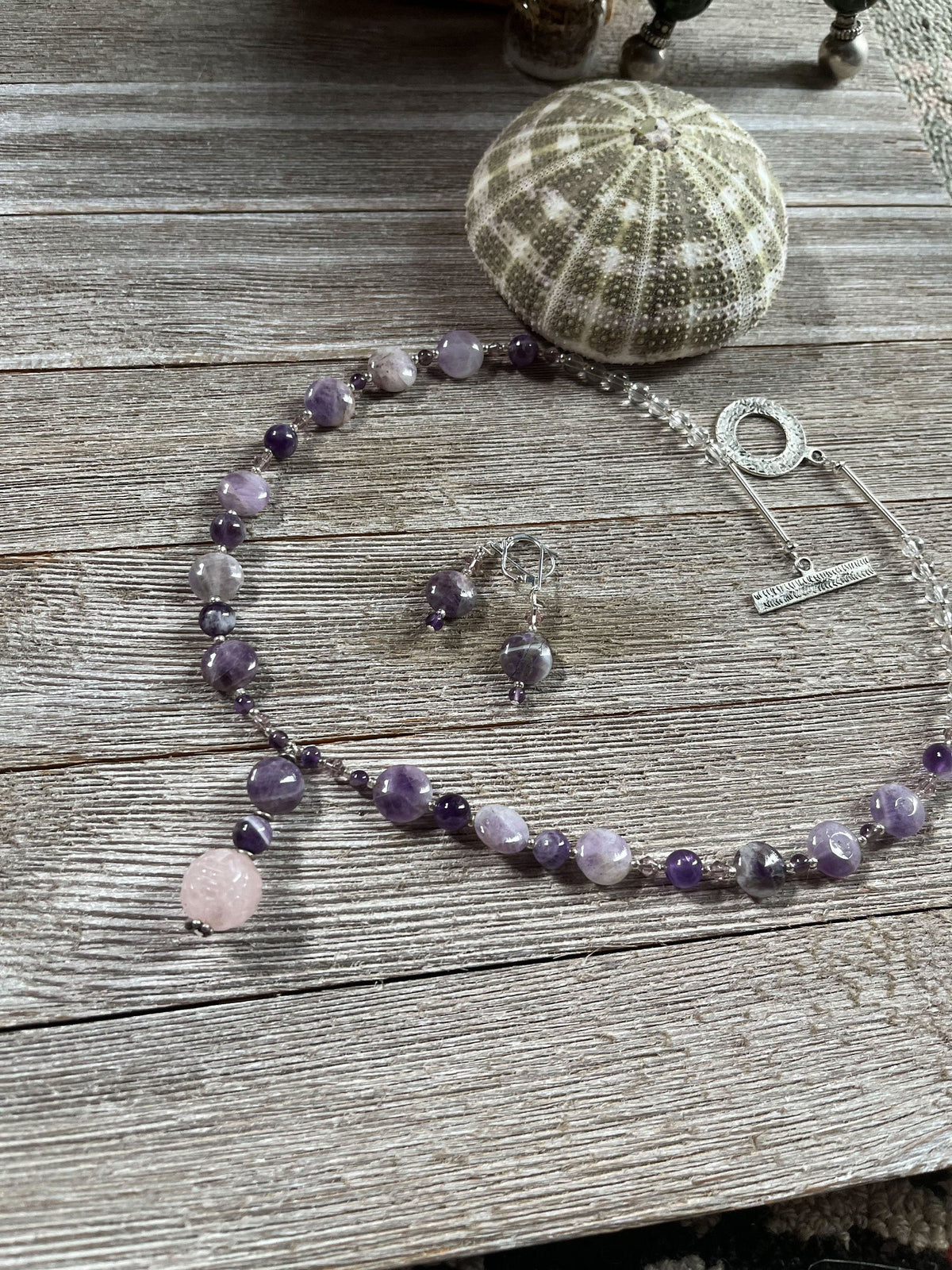 The Artisan Designed Jewelry Set features an amethyst and rose quartz necklace with sterling silver earrings, USA handcrafted, displayed on a rustic wooden surface next to a green sea urchin shell.