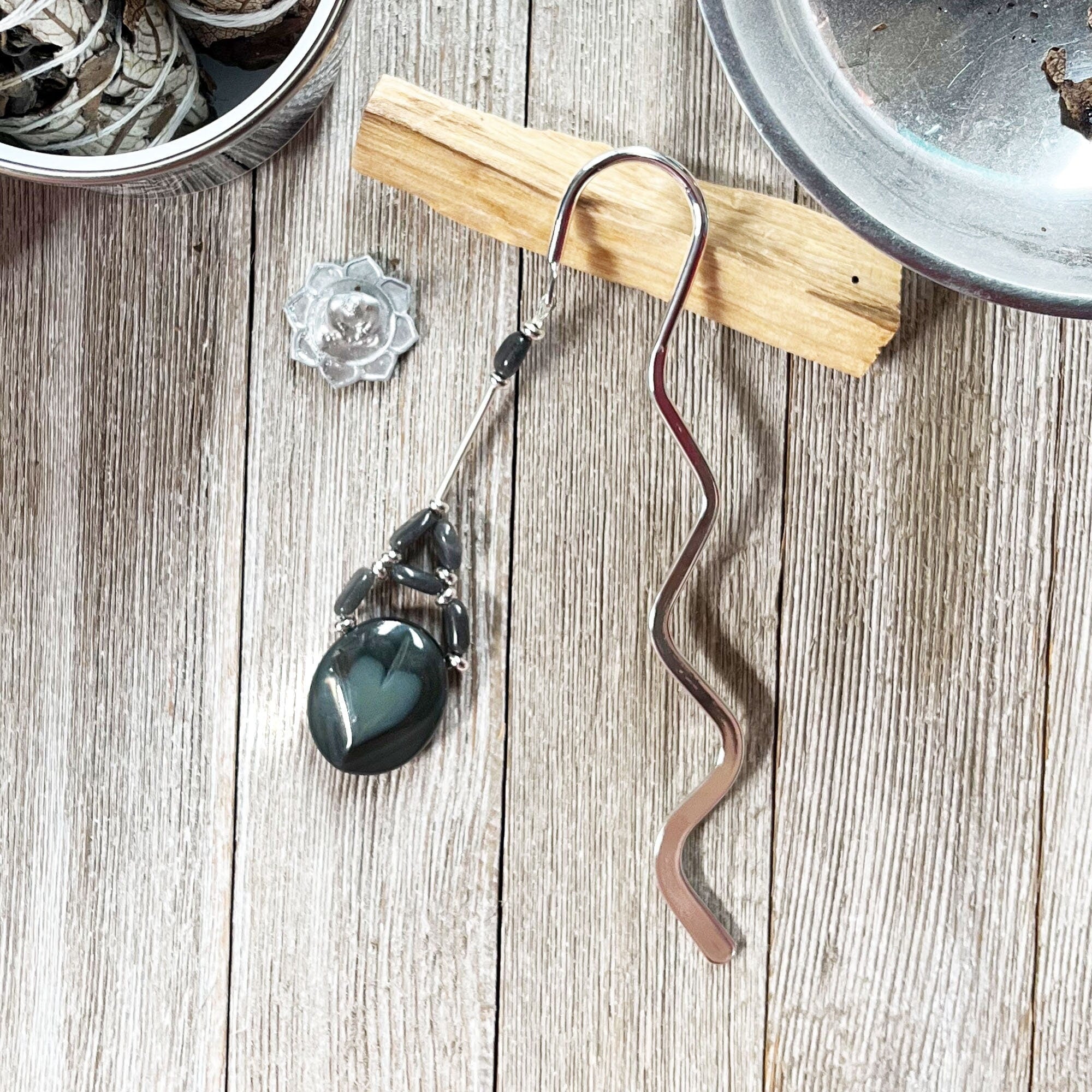 The Labradorite Black Heart Stone Bookmark features a wavy design and black gemstone charm, hanging from a wooden holder on a light surface near dried herbs—a thoughtful Wiccan gift.