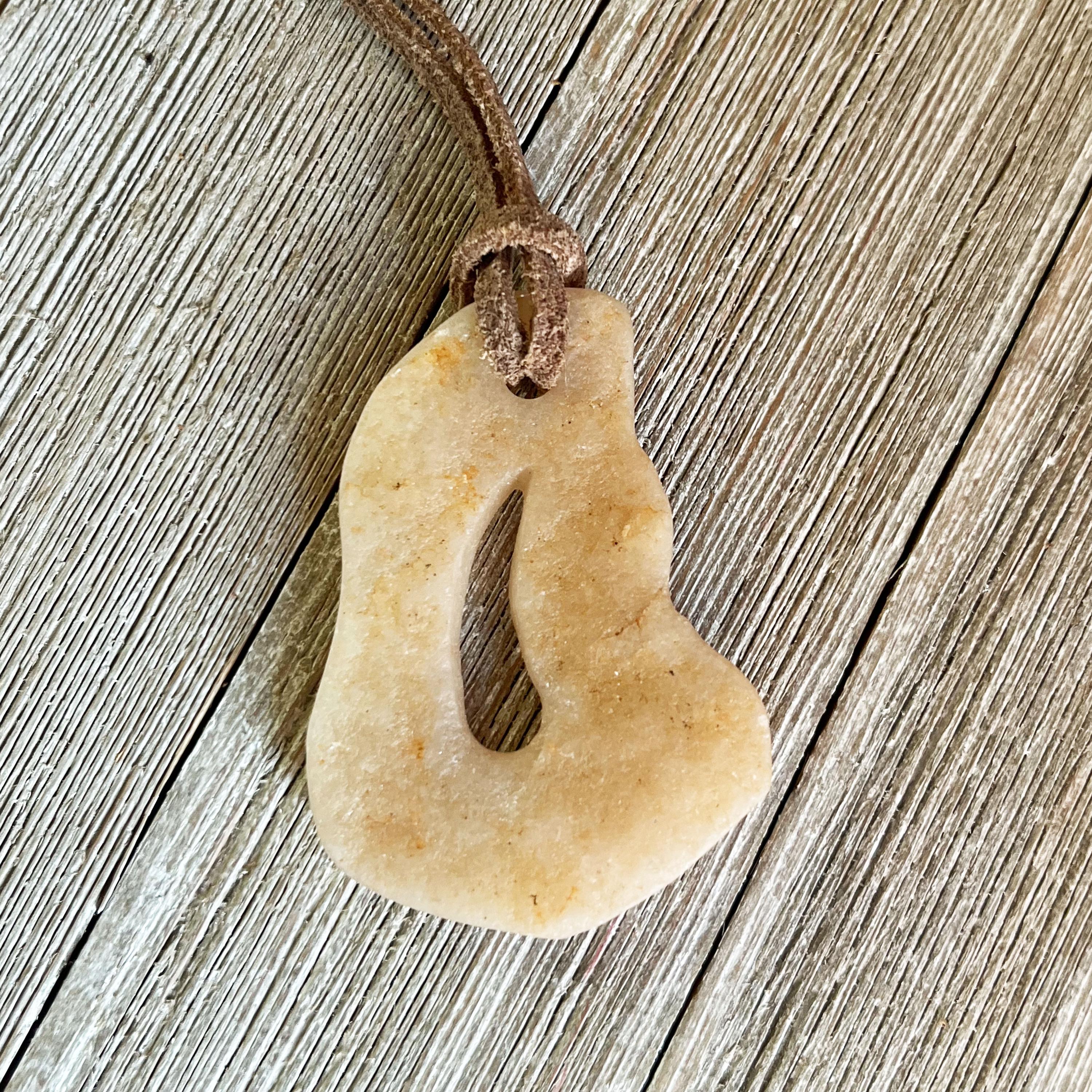 The Stone Pendant Necklace features a beige, freeform jasper pendant with a central cut-out on an adjustable leather cord, shown resting on rustic wood with visible grain.