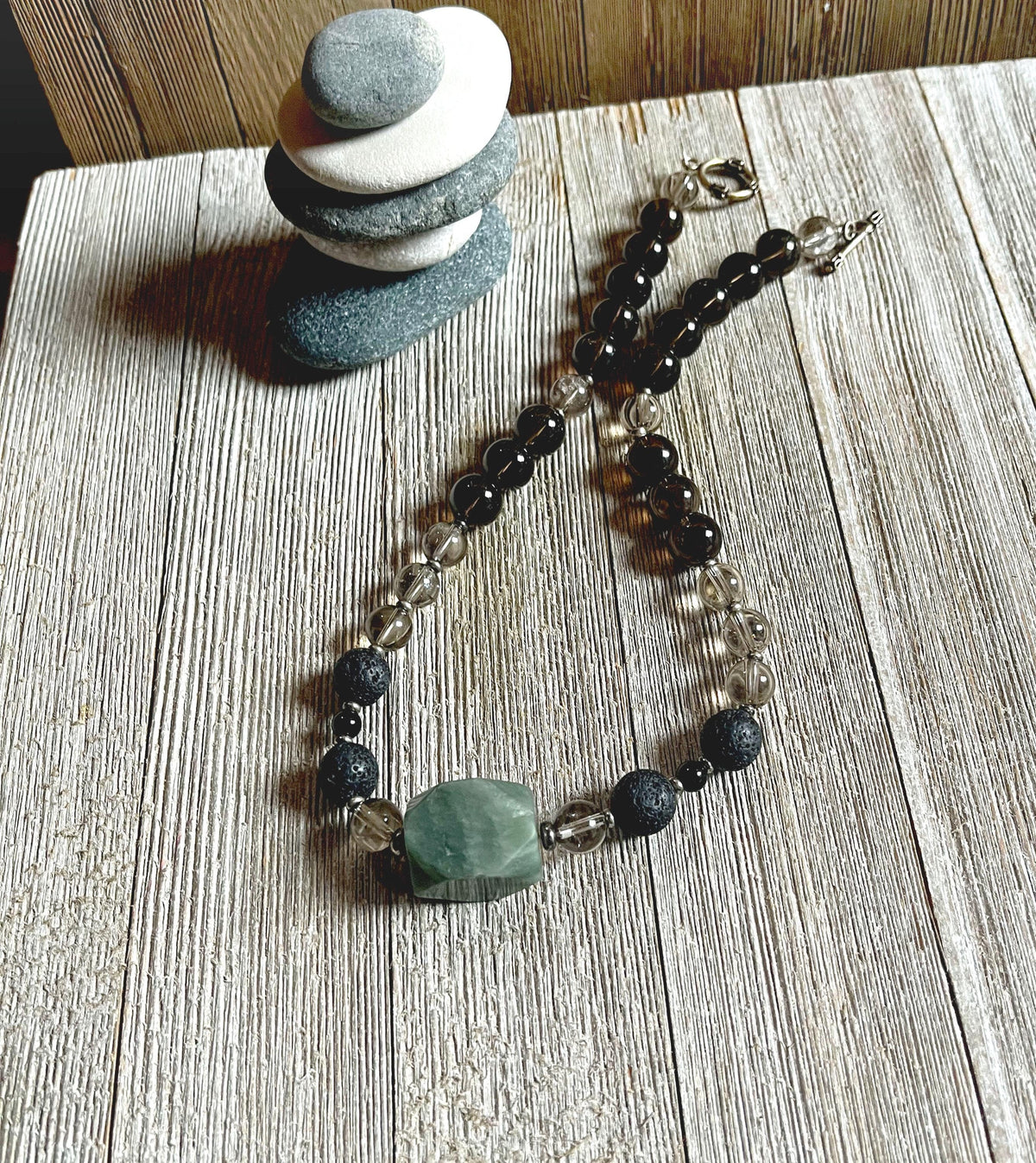 The Breciated Jasper Green Stone Necklace—an artisan-made essential oil diffuser with therapeutic lava beads and a minimalist design—rests on a wooden surface beside a small stack of balanced stones, making it a unique gift for him.