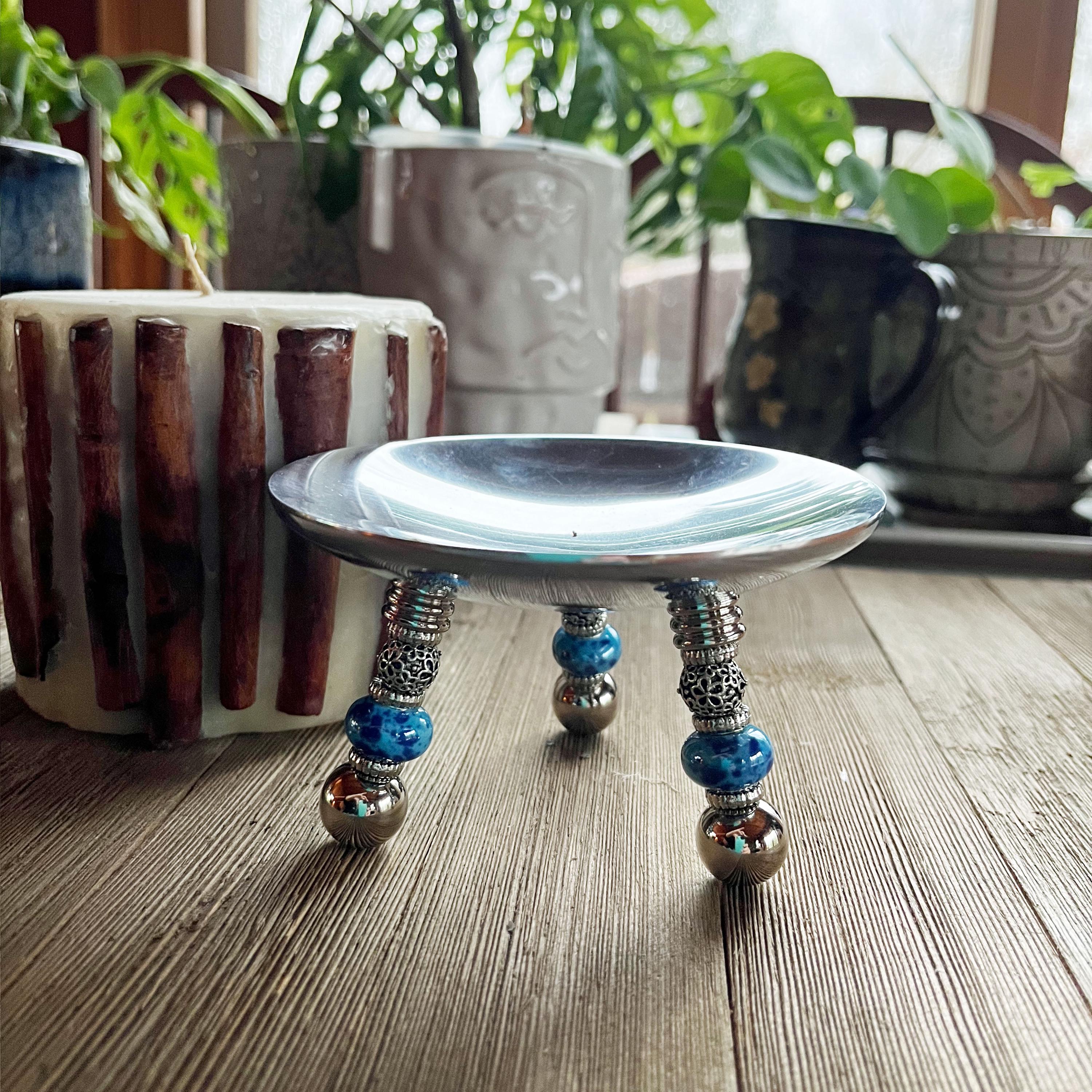 The Candle Holder—an artistic stainless steel piece with blue beaded legs—rests on a wooden surface as a unique table centerpiece, surrounded by potted plants and mugs, illuminated by natural light.