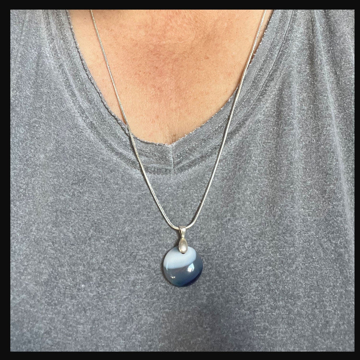 A person wears the Blue Agate Reflection Eye Necklace: Minimalist Zen Pendant—handcrafted in the USA—over a heather gray v-neck tee, adding a subtle, stylish accent with its round blue and white striped design.