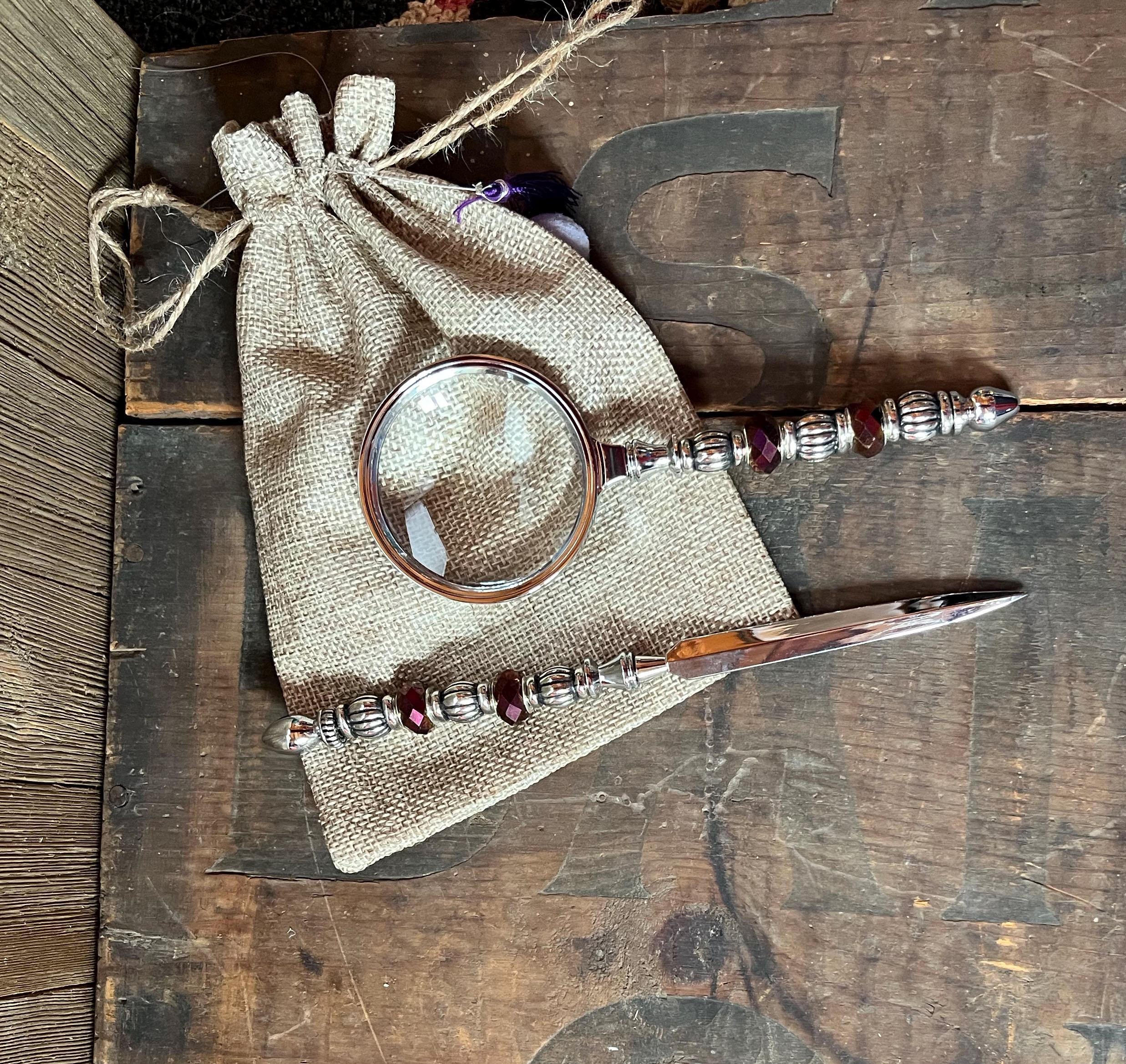The Purple Magnifier and Letter Opener - Beaded Desk Set features ornate silver and red handles, displayed on a burlap pouch atop a rustic wooden surface.
