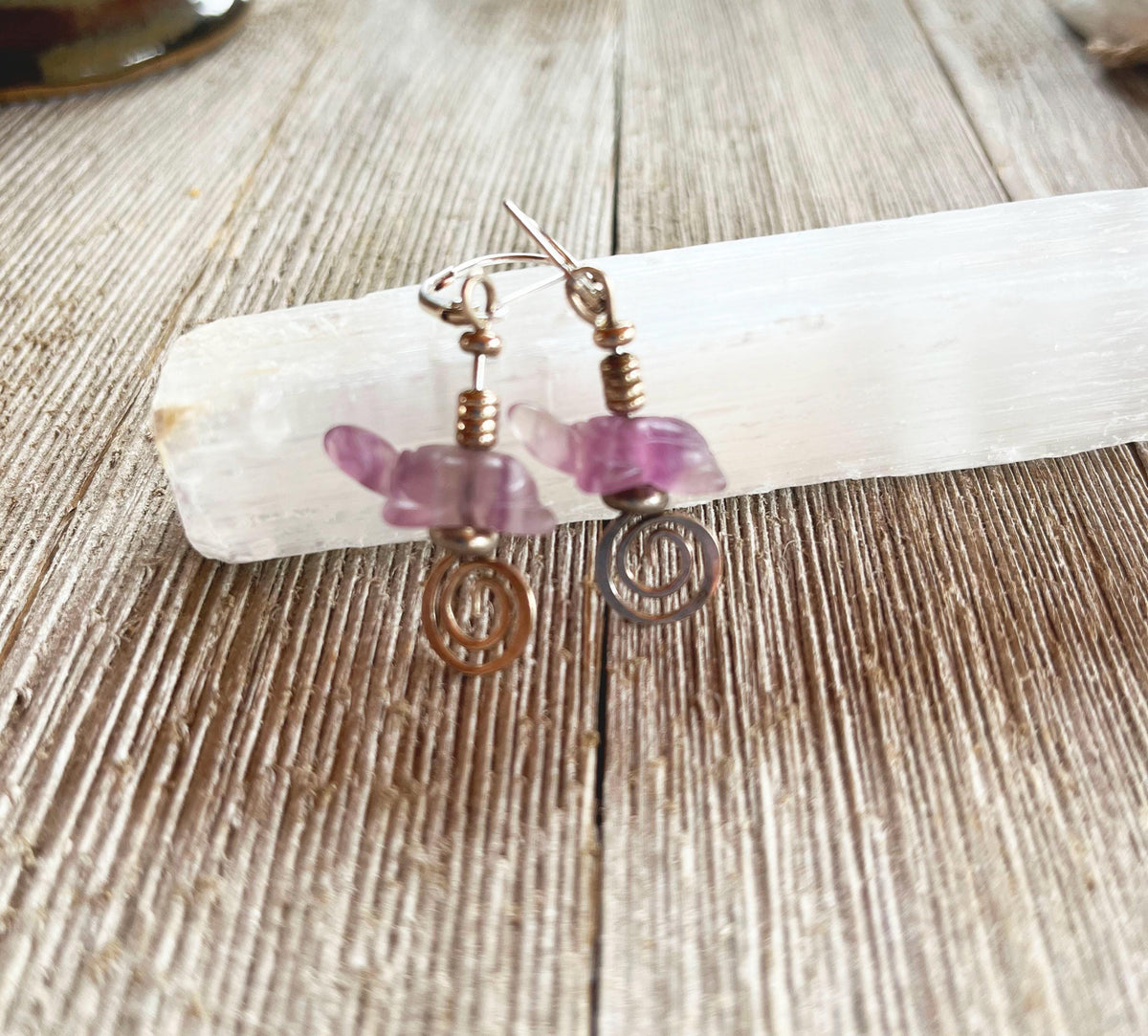 Turtle Earrings Amethyst Carved with Silver Swirl Detail are shown hanging on a white crystal atop a rustic wooden surface—a beautiful, nature-inspired jewelry piece for animal lovers.