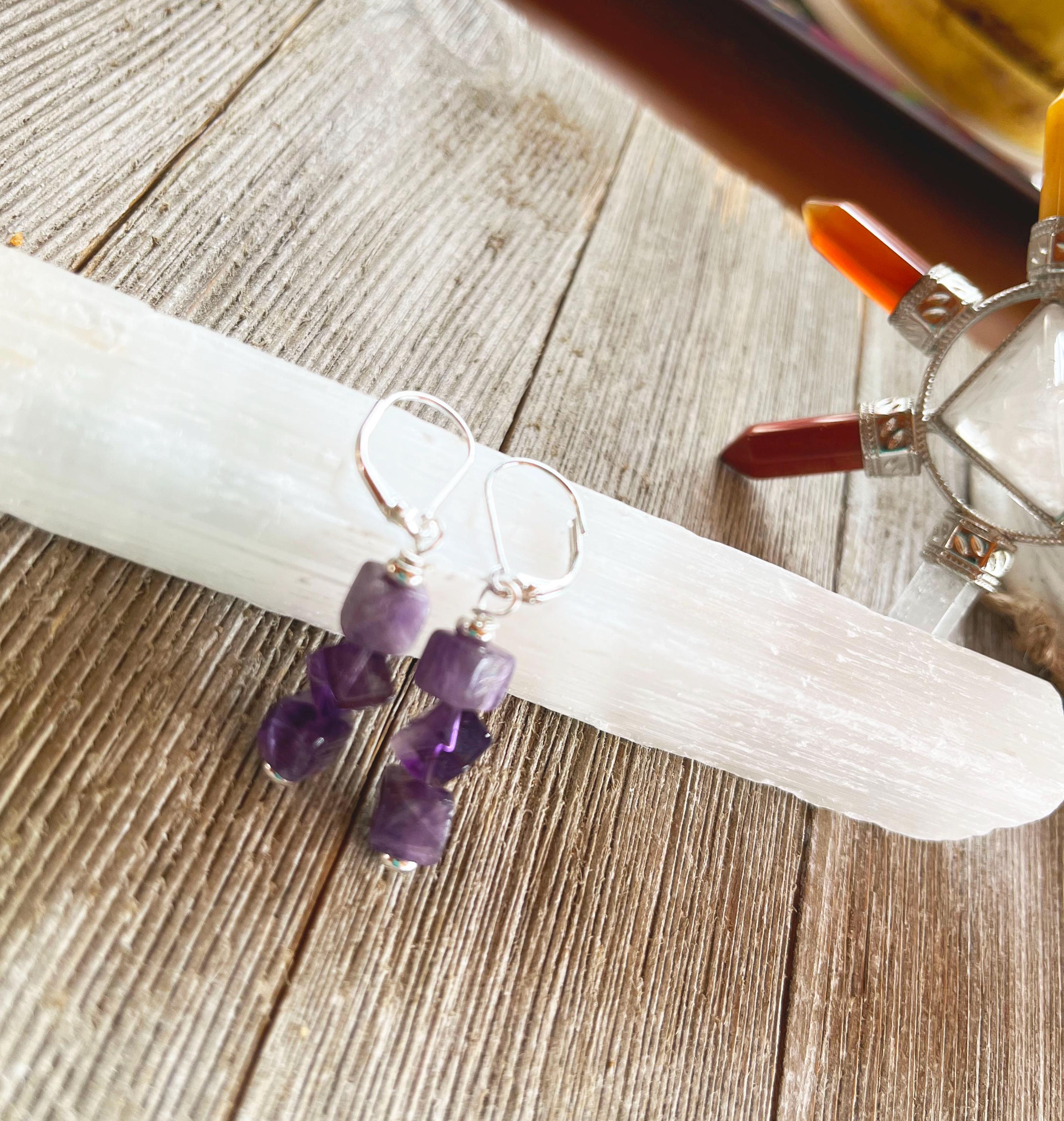 Spiritual Jewelry in Purple Stone Amethyst Cube Earrings—minimalist and handmade in the USA—rest on a white crystal wand atop wood, with a vibrant crystal holder featuring colorful points seen in the background.