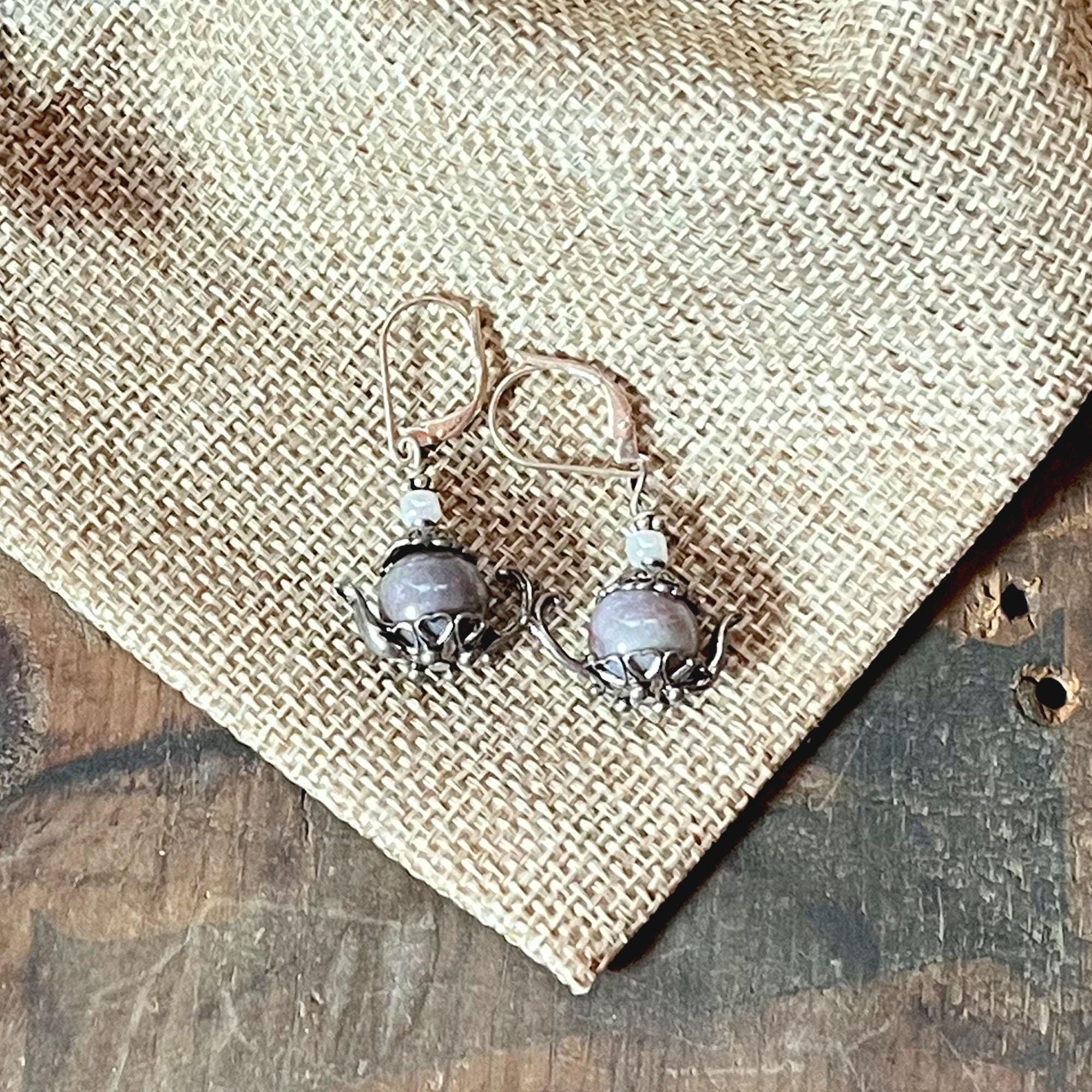 Sterling Silver Teapot Earrings feature round lavender jasper stones and whimsical design, displayed on burlap fabric over a wooden surface.