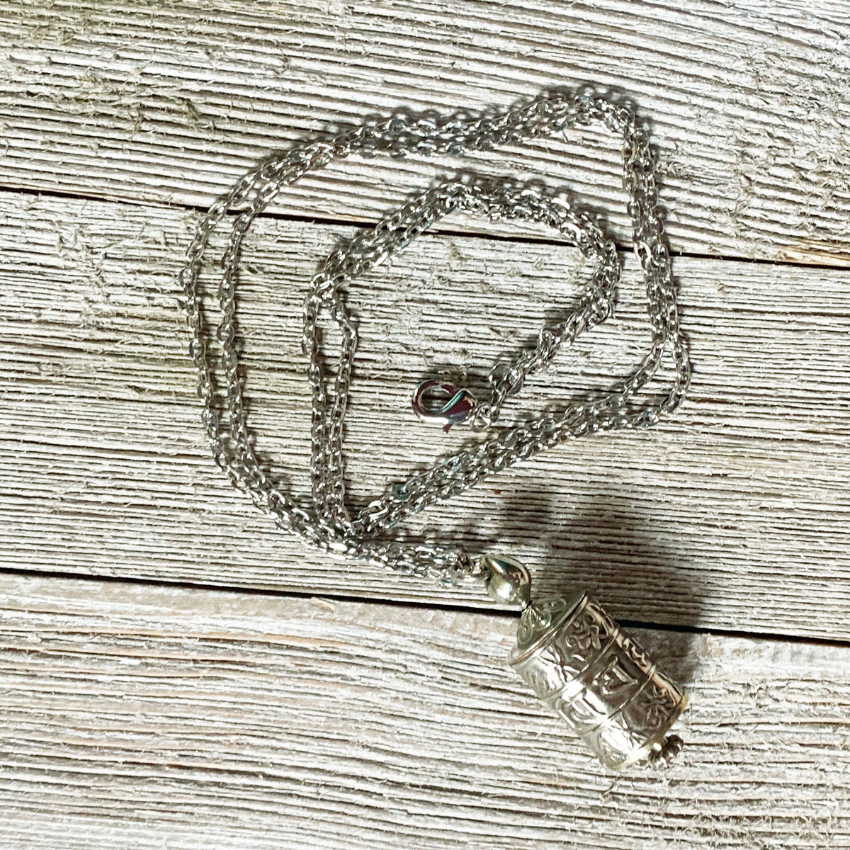 The Prayer Wheel Necklace features a silver Zen Mantra Pendant with intricate patterns, designed for mindful meditation, shown resting on a textured wooden surface.