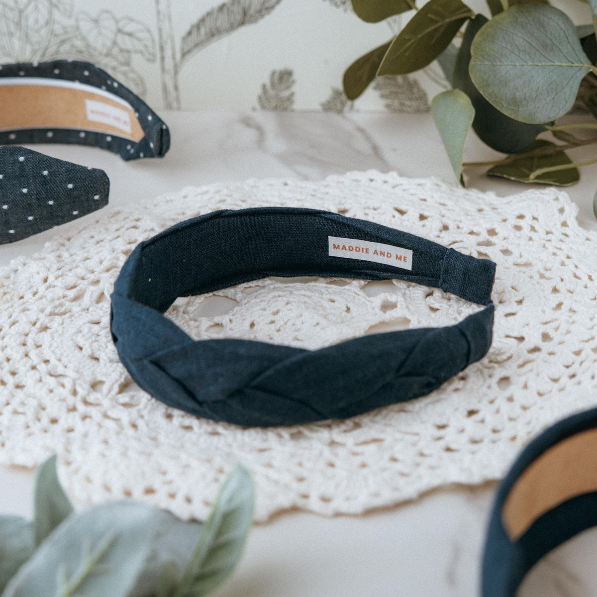 A dark navy blue braided headband with a label that reads "MADDIE AND ME" is displayed on a white lace doily. Other headbands with white polka dots and a tan interior are also visible. The background includes greenery and a patterned wall.