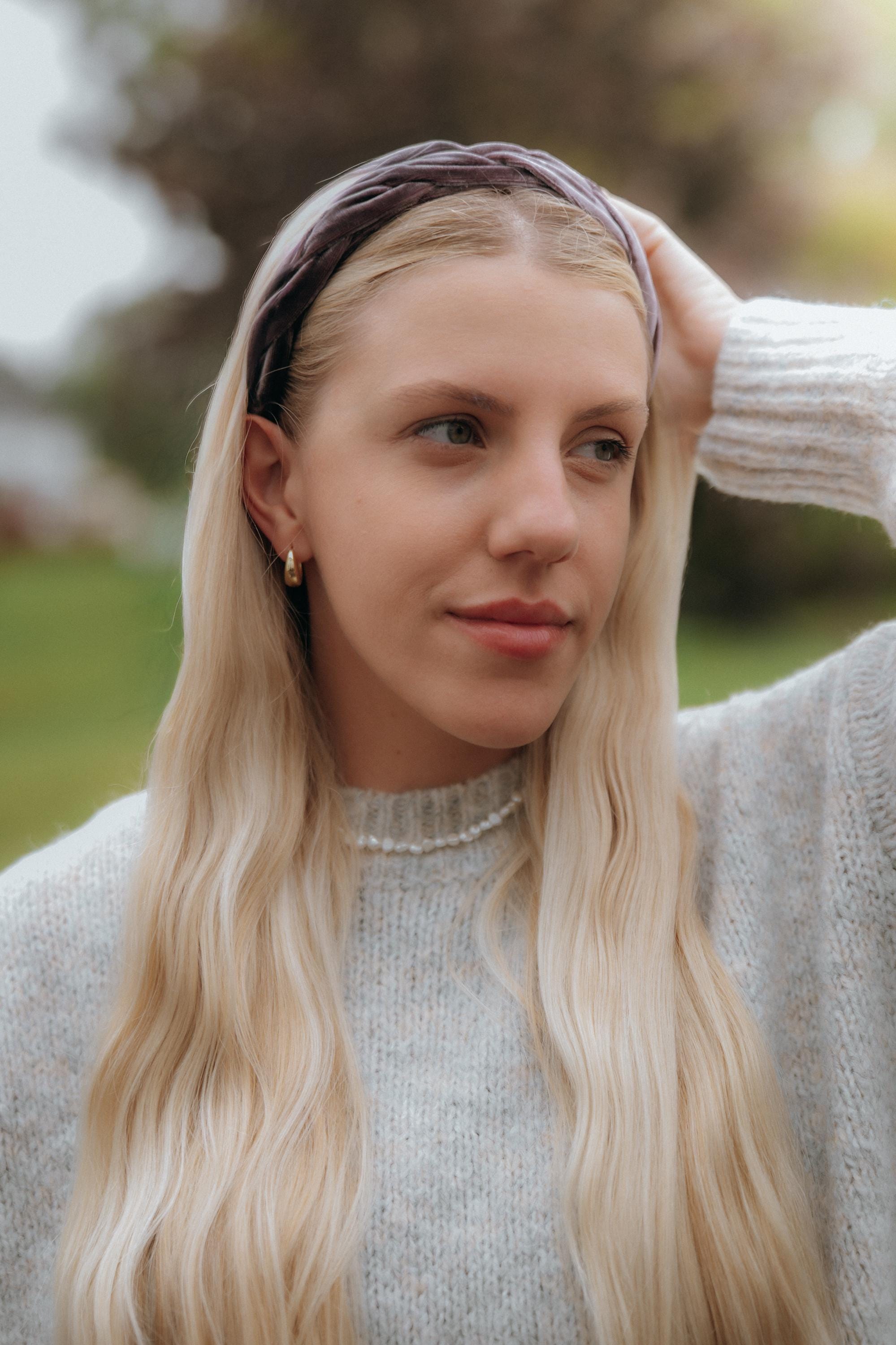 May include: A woman with long, blonde hair wearing a gray sweater, a pearl necklace, and a mauve velvet headband. She is also wearing gold hoop earrings. The background is blurred with green and brown tones.