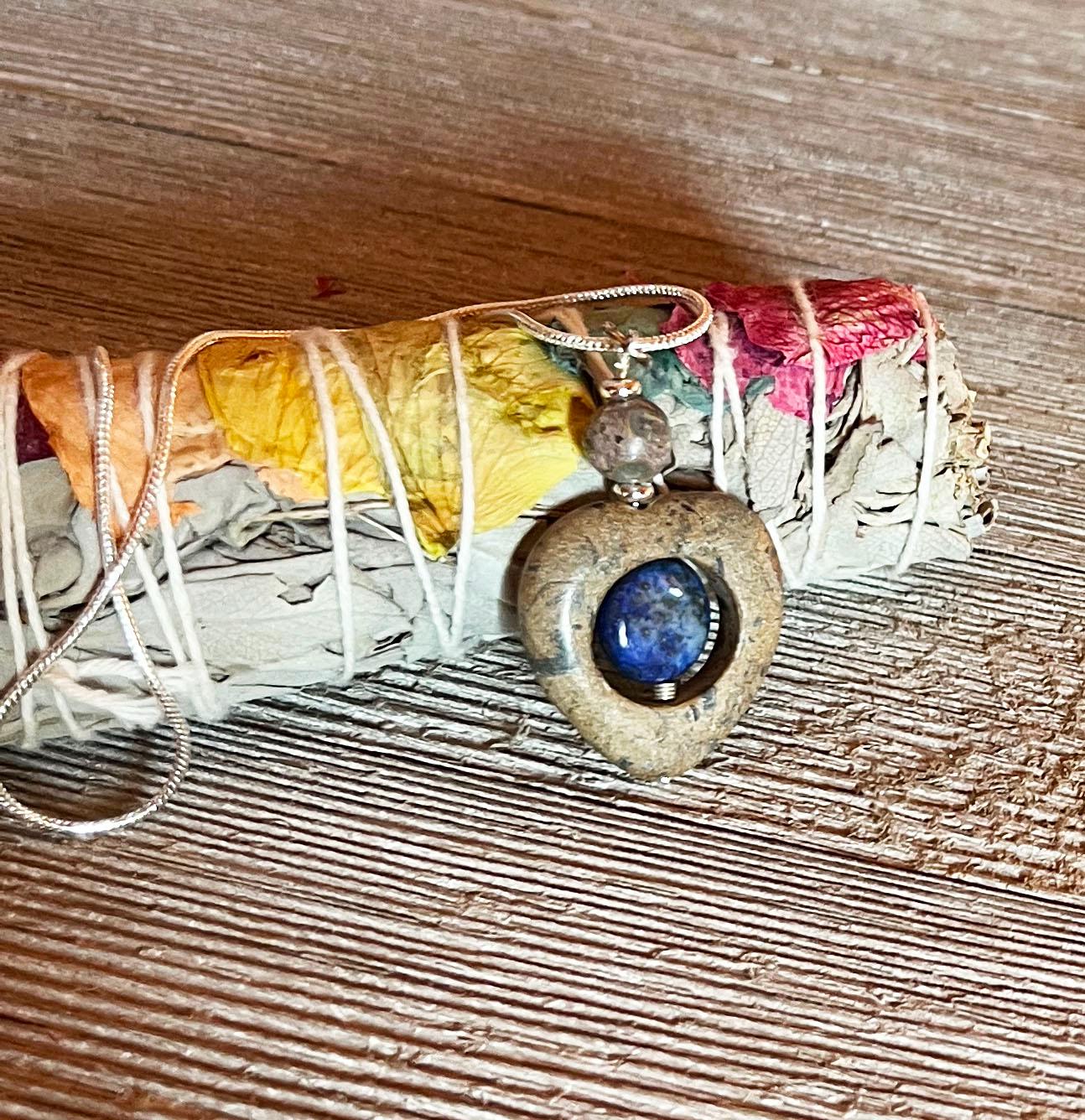 The Lapis Lazuli Spinner Pendant—featuring a blue jasper stone—rests on dried sage and petals, wrapped in white string atop textured wood, making this necklace the perfect calming fidget jewelry.