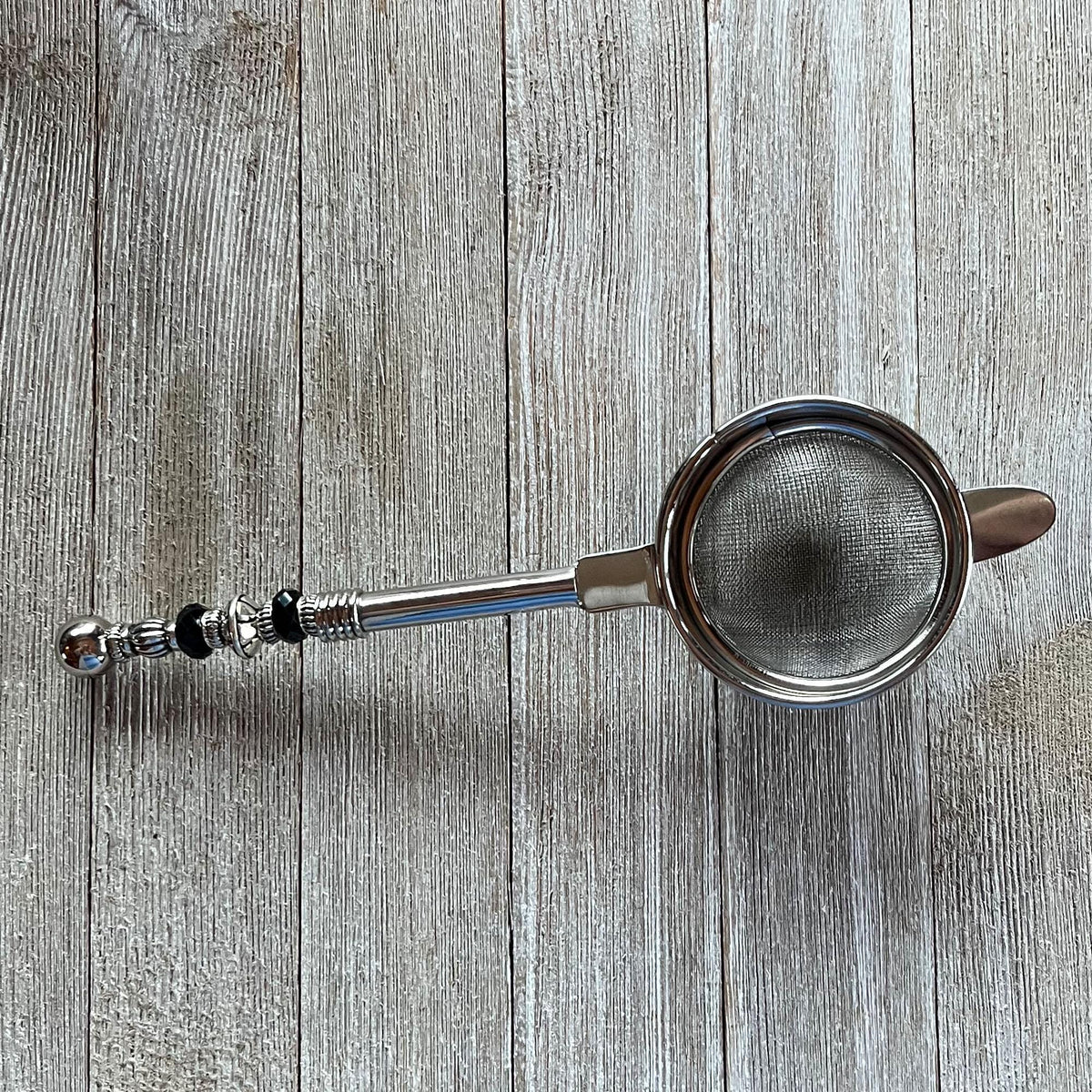 The Beaded Tea Strainer – Handcrafted Customizable Handle (9” Length), adorned with black and silver accents, rests elegantly on a light wooden surface—an ideal gift for tea lovers.