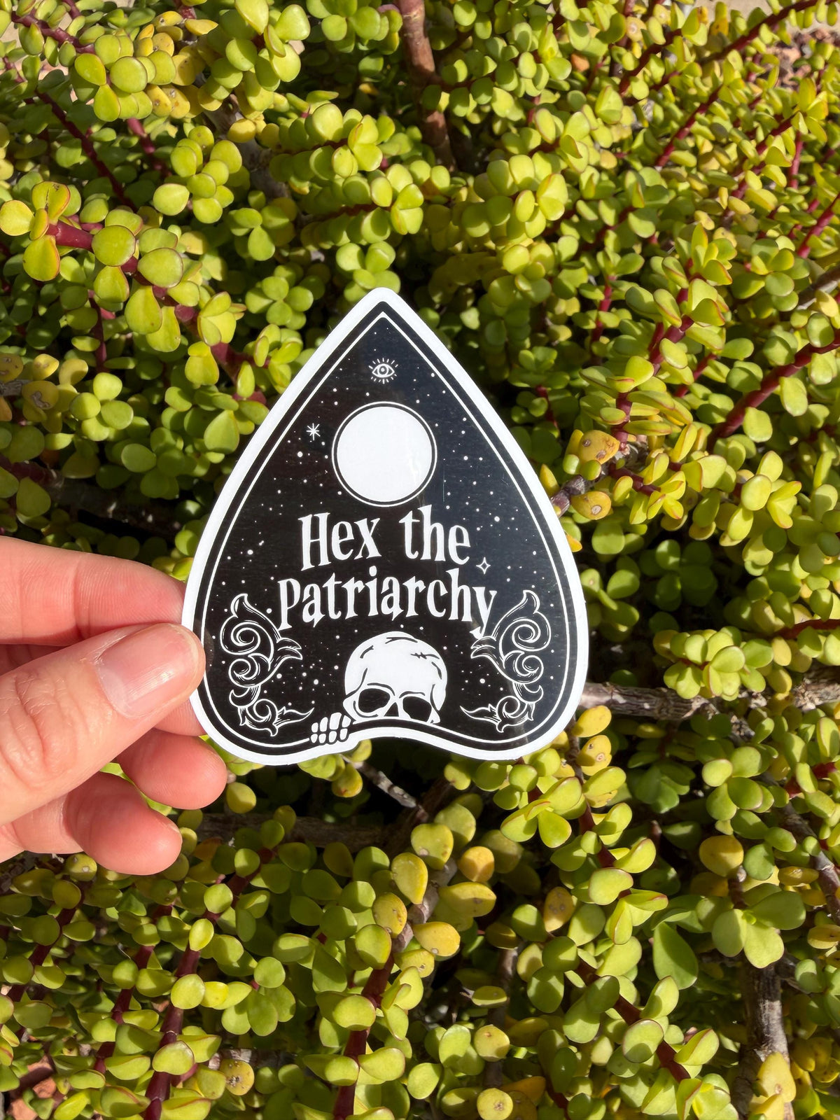 May include: A black and white sticker in the shape of a planchette, with the words "Hex the Patriarchy" in white script. The sticker features a skull, moon, and stars design. The sticker is held in front of a background of green leaves.